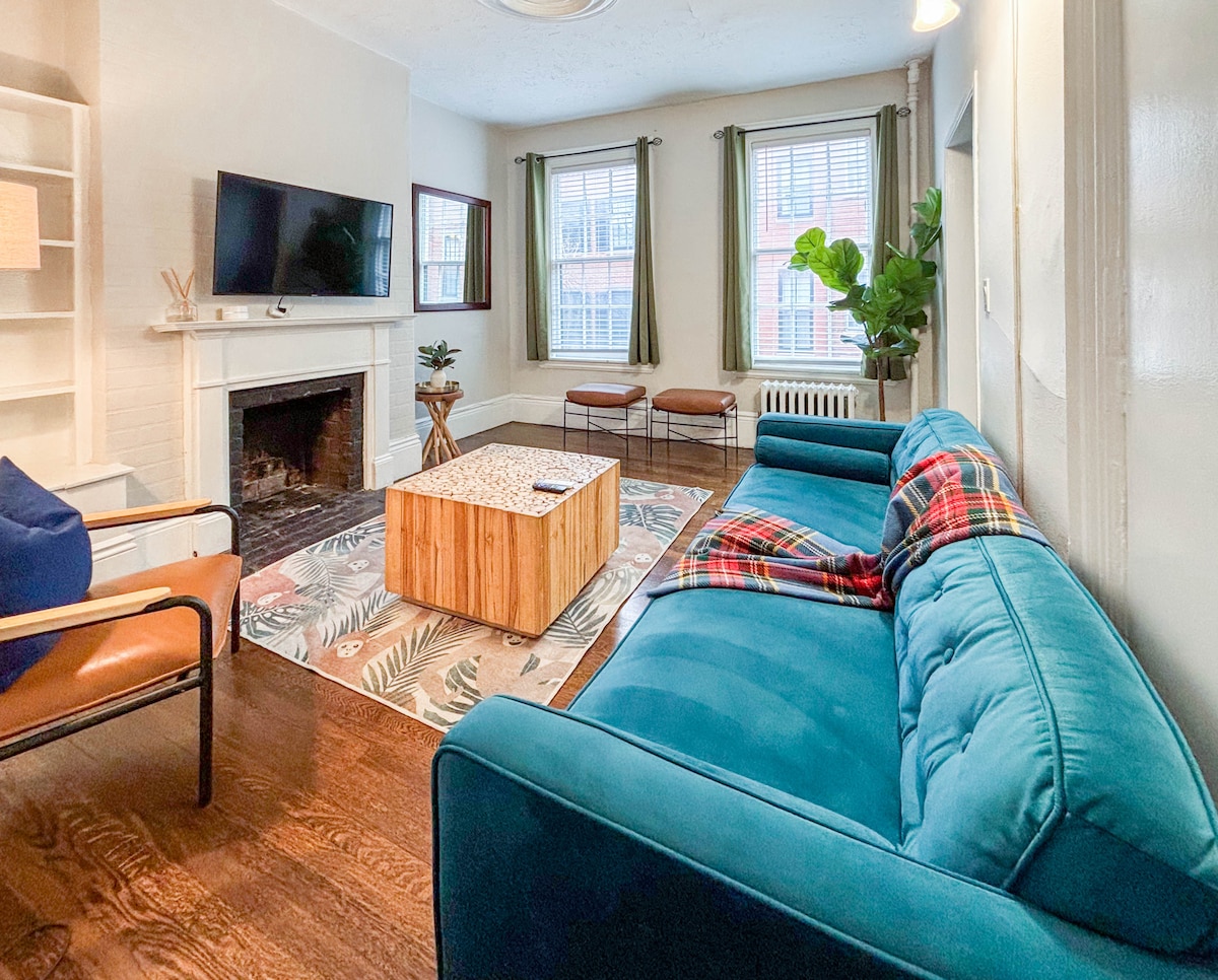 The living room features a cozy seating area with a blue sofa and wooden coffee table positioned in front of a flat-screen TV. Natural light filters through the windows adorned with light curtains, while indoor plants add a touch of greenery to the space.