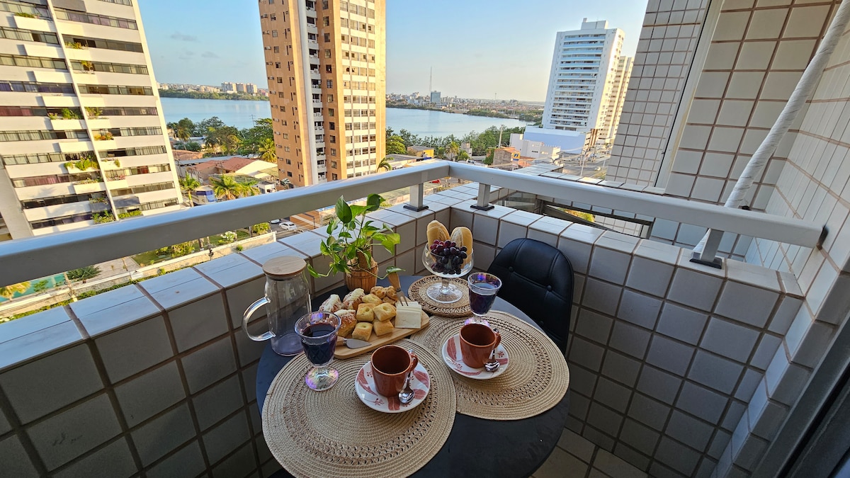 A balcony table is set for two, featuring plates of cookies and drinks. A plant adds a touch of greenery, while the waterfront view showcases distant city buildings and the shimmering lagoon. The tile design complements the urban environment.