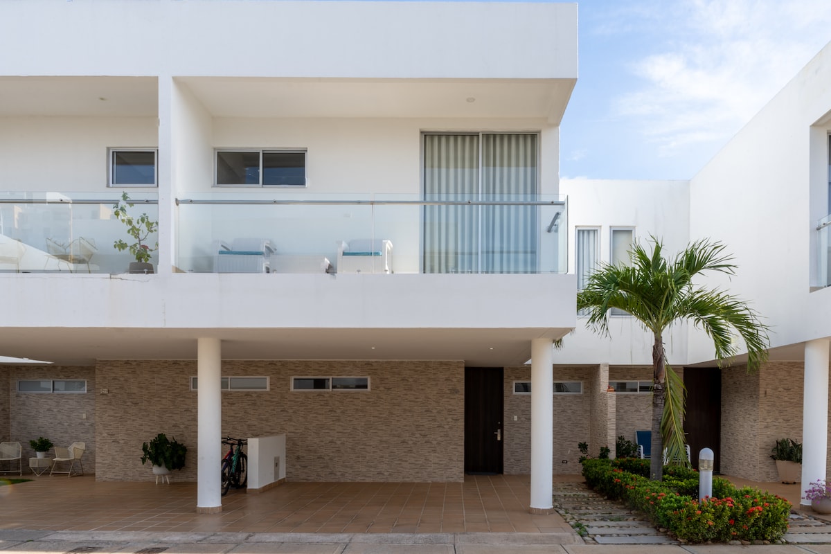 The exterior of a modern property features a clean design with a white facade, large glass windows, and a balcony. Potted plants are positioned on the balcony. A palm tree adds greenery to the entrance, with a paved pathway leading to the doors.