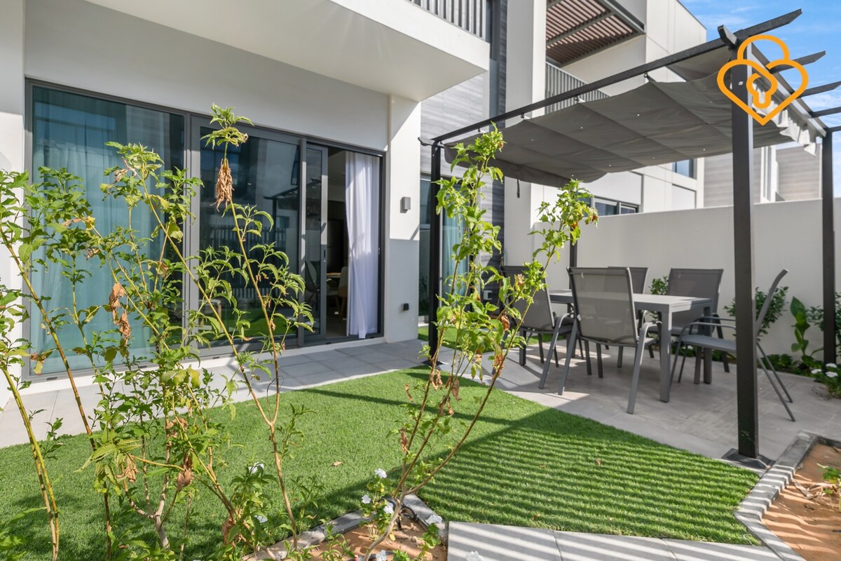 An outdoor space features a covered pergola with a dining table and chairs, surrounded by green landscaping. The area is designed for relaxation and enjoyment, with sliding glass doors providing access to the interior of the townhouse.