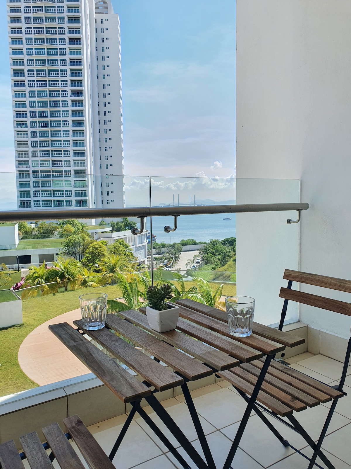 A small balcony features a wooden table and two folding chairs, providing a space for relaxation. Views of the coastline and greenery can be seen in the background, while tall buildings rise in the distance against a clear blue sky.