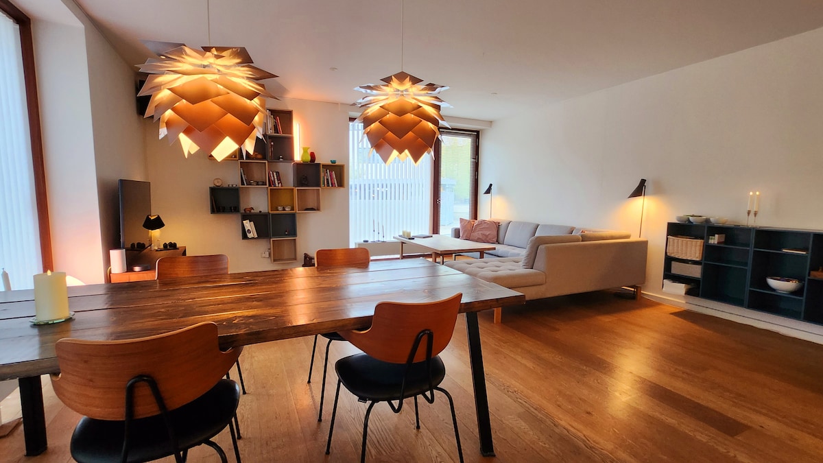 An open living and dining area is featured, with a large wooden table surrounded by black chairs. Two contemporary light fixtures hang above the table. A comfortable sofa is positioned beside a window, providing natural light, while bookshelves and decorative elements create an inviting environment.