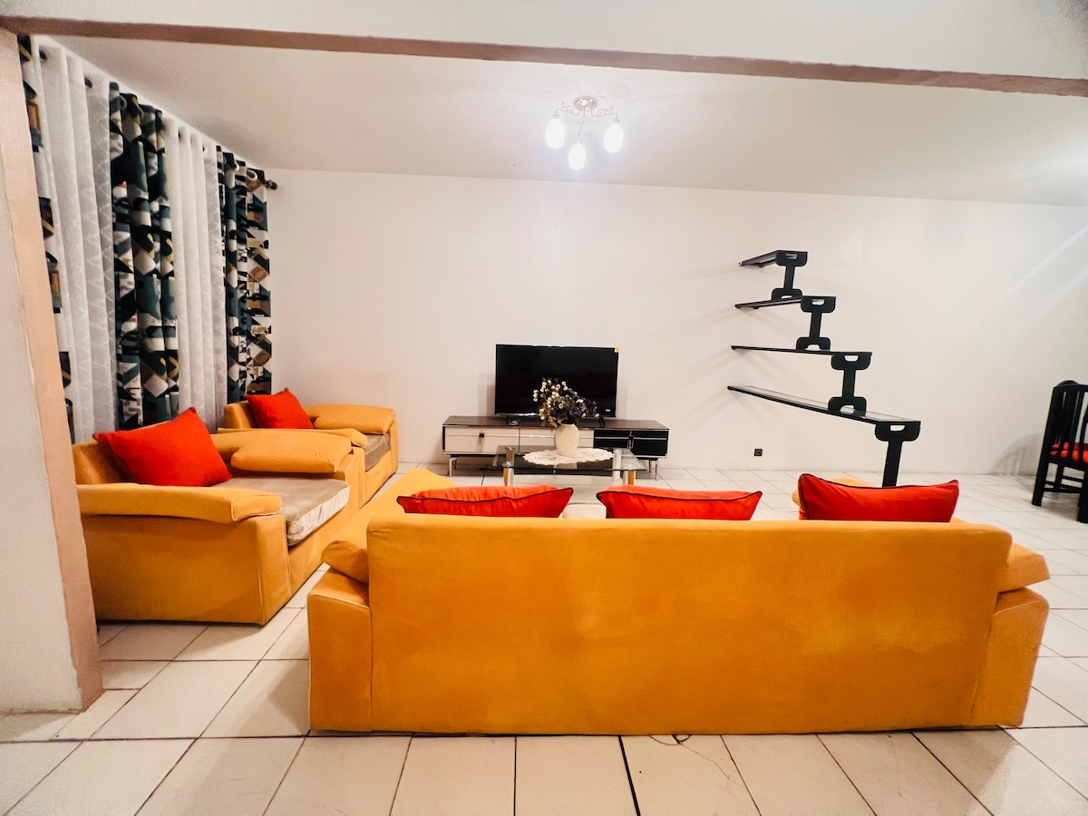 The living room features a bright, open layout with yellow sofas adorned with contrasting red cushions. A television is situated on a sleek, modern console. Large windows are adorned with patterned curtains, while a unique staircase leads to an upper level.