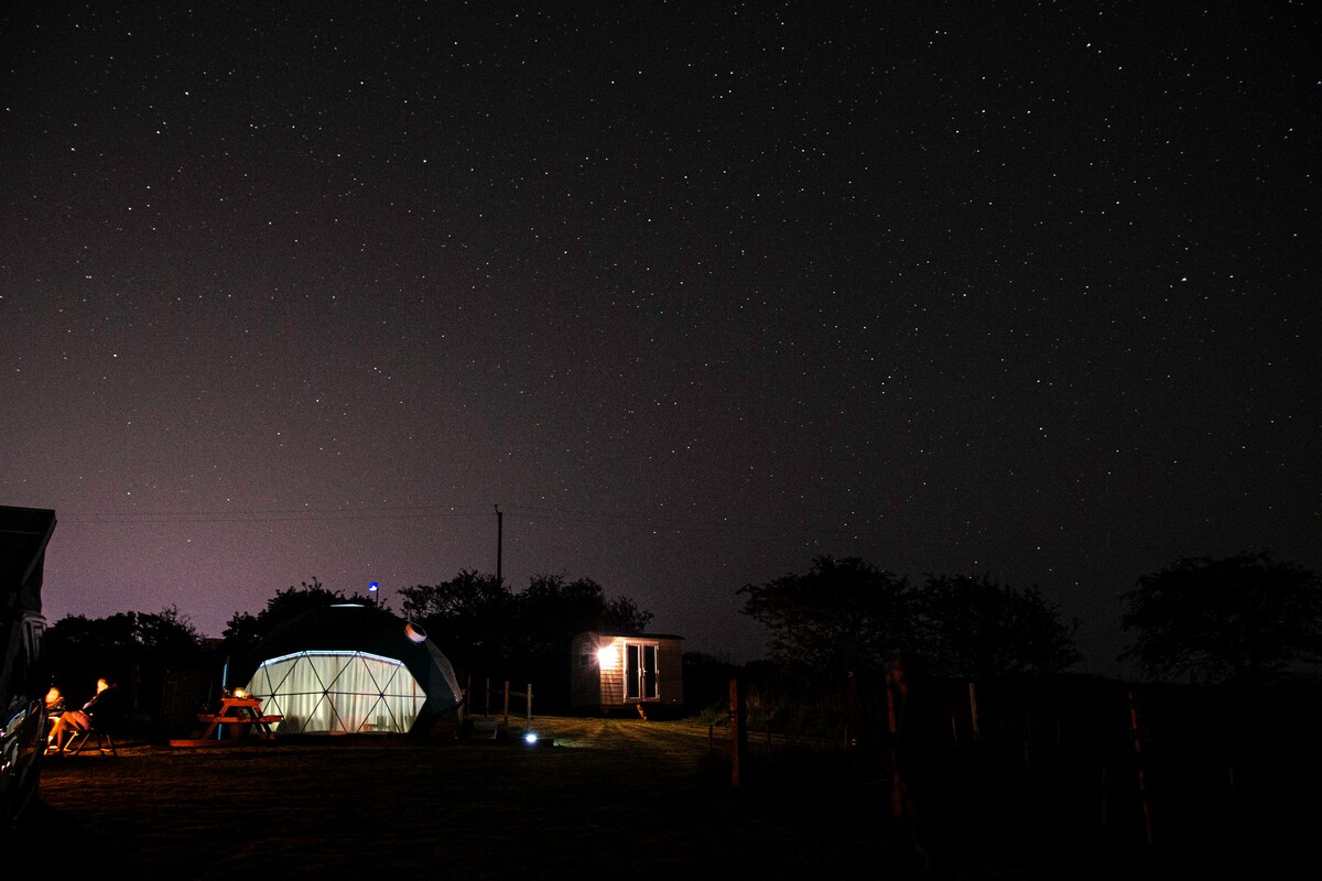 A geodesic dome stands under a starry sky, surrounded by a peaceful landscape. A shepherd's hut restroom is visible nearby, with soft lighting illuminating the dome. A picnic table is positioned on the lawn, inviting relaxation in the tranquil evening setting.