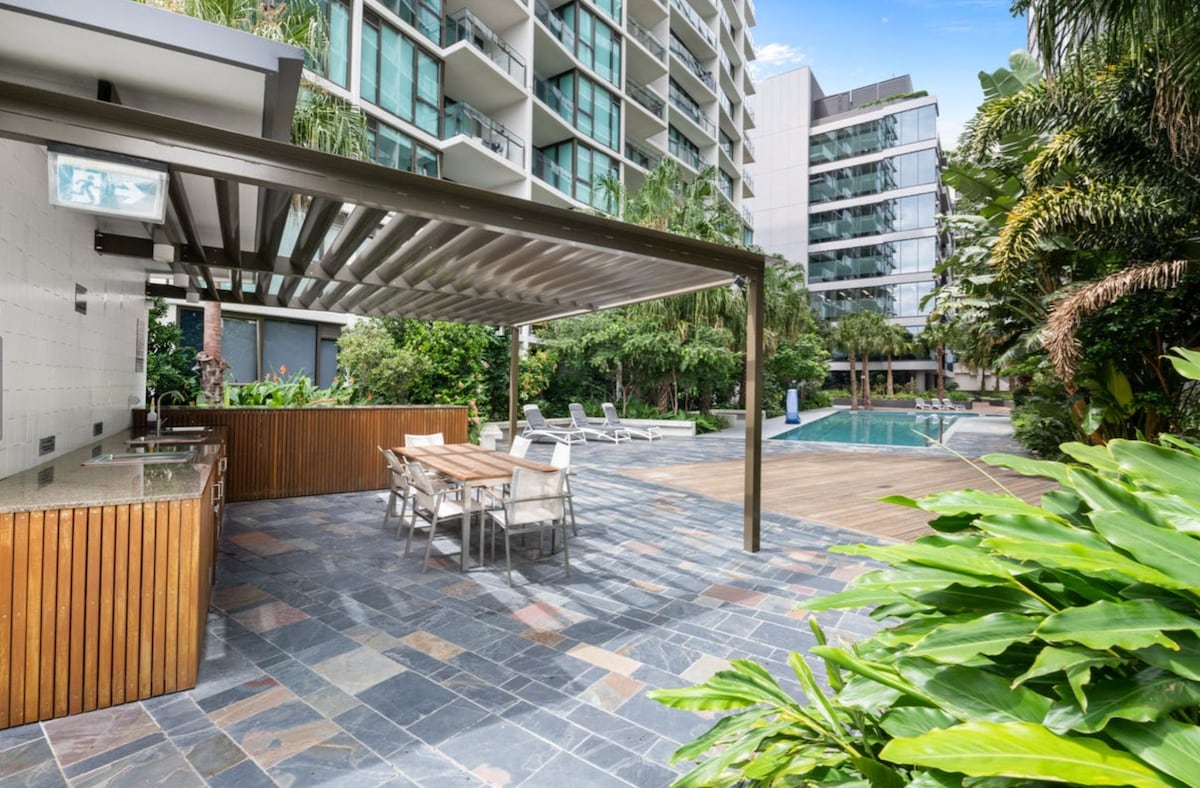 An outdoor area features a covered BBQ space with a long dining table and chairs. A stone pathway leads to a sparkling pool surrounded by lush greenery and lounge chairs, creating a relaxing environment for guests.