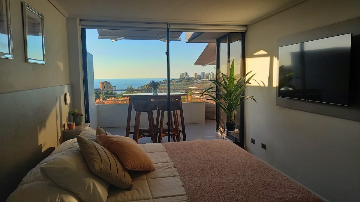 A bright sleeping area features a spacious bed adorned with soft pillows. Large glass windows provide a view of the coastline and cityscape beyond. A minimalist television hangs on the wall, while a small plant adds a touch of greenery to the space.