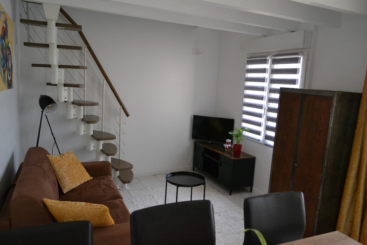 A bright living area features a comfortable brown sofa adorned with yellow cushions. A small black side table and a television stand are present. Large windows with horizontal blinds allow natural light to fill the space, while a staircase leads to an upper level.