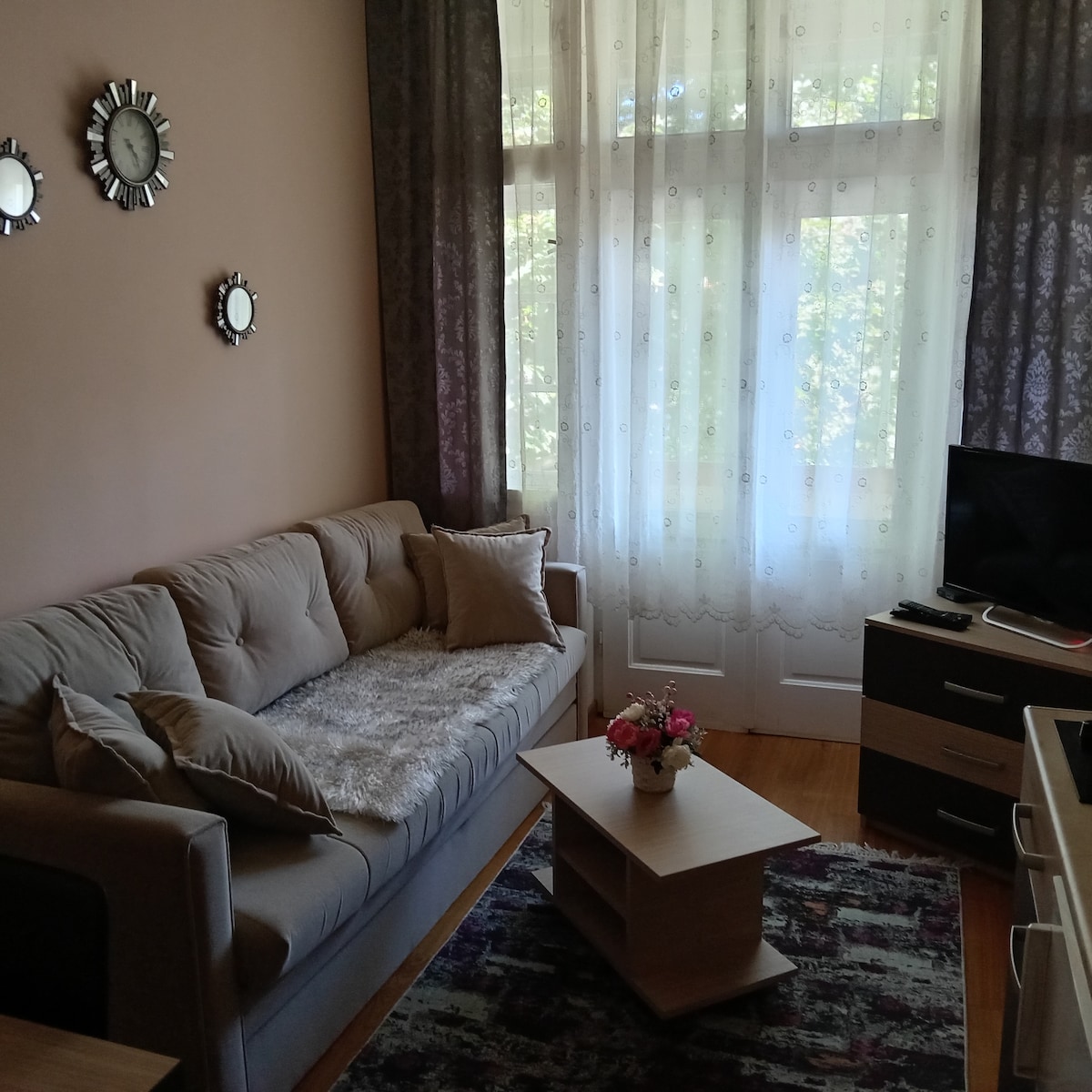 A comfortable seating area is featured, with a light-colored sofa adorned with decorative pillows and a soft throw. A small coffee table is positioned in front, and natural light filters through the sheer curtains of an adjacent window.