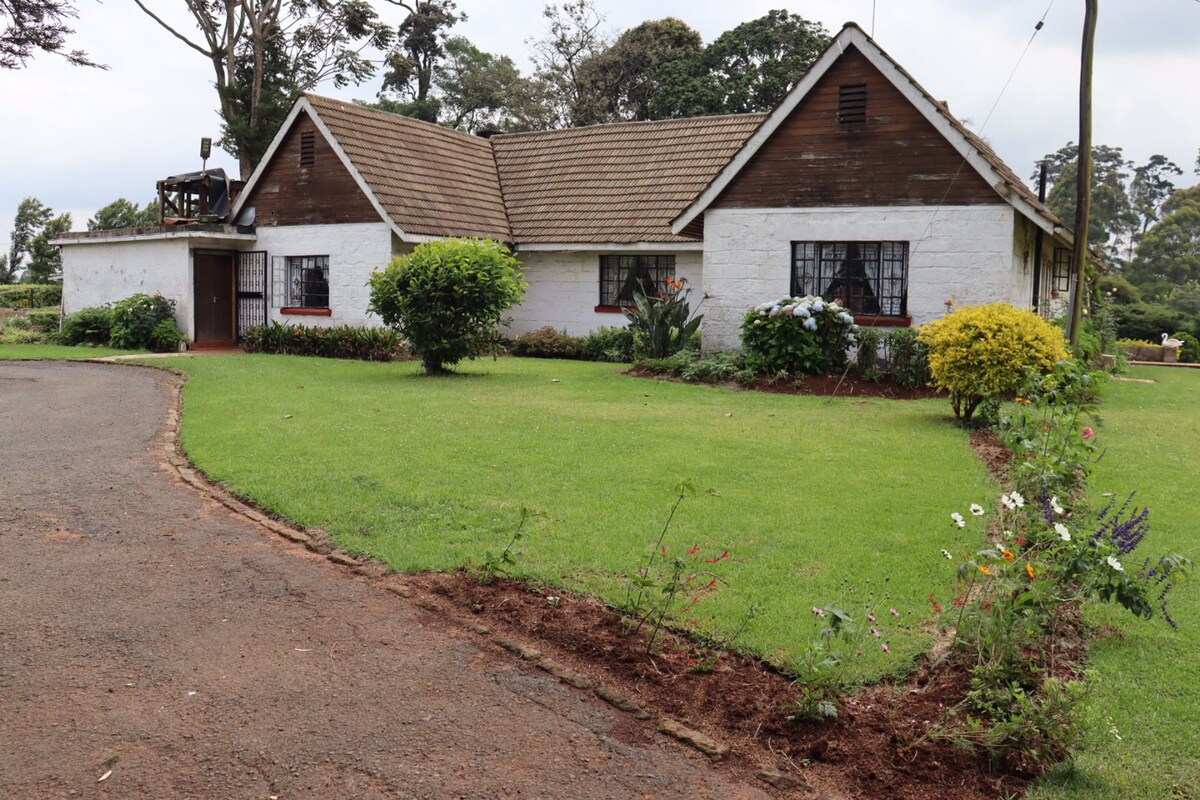 The exterior of a charming house is framed by well-maintained green lawns and colorful flower beds. The structure features a mix of white and wooden elements, with large windows enhancing natural light. Surrounded by trees, the serene setting provides a peaceful ambiance.