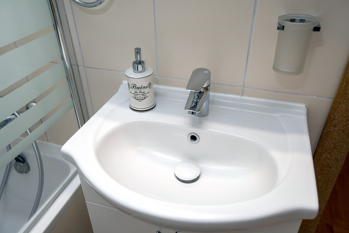 A well-maintained bathroom sink features a smooth, rounded basin and a modern faucet. A decorative soap dispenser adds a touch of elegance, and a wall-mounted container is visible nearby. Soft lighting enhances the clean, tiled surroundings.