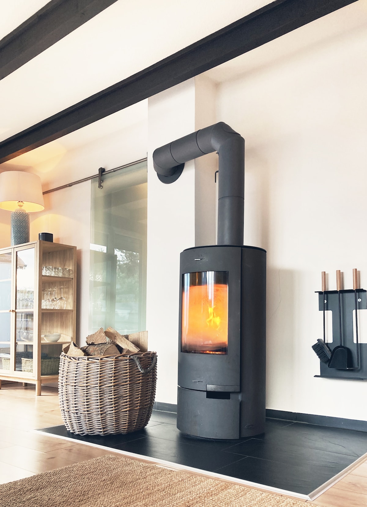 A modern wood stove stands against a neutral wall, featuring a glass front that reveals flickering flames. A woven basket filled with firewood rests nearby, while a rack for fire tools is fixed to the wall beside the stove.