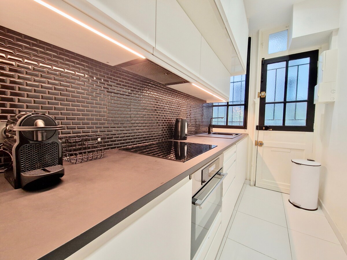 A modern kitchen features sleek cabinetry with a dark textured backsplash. Under-cabinet lighting illuminates the countertop, where a coffee machine and kettle are visible. Large windows allow natural light to fill the space, while the door leads to the rest of the apartment.
