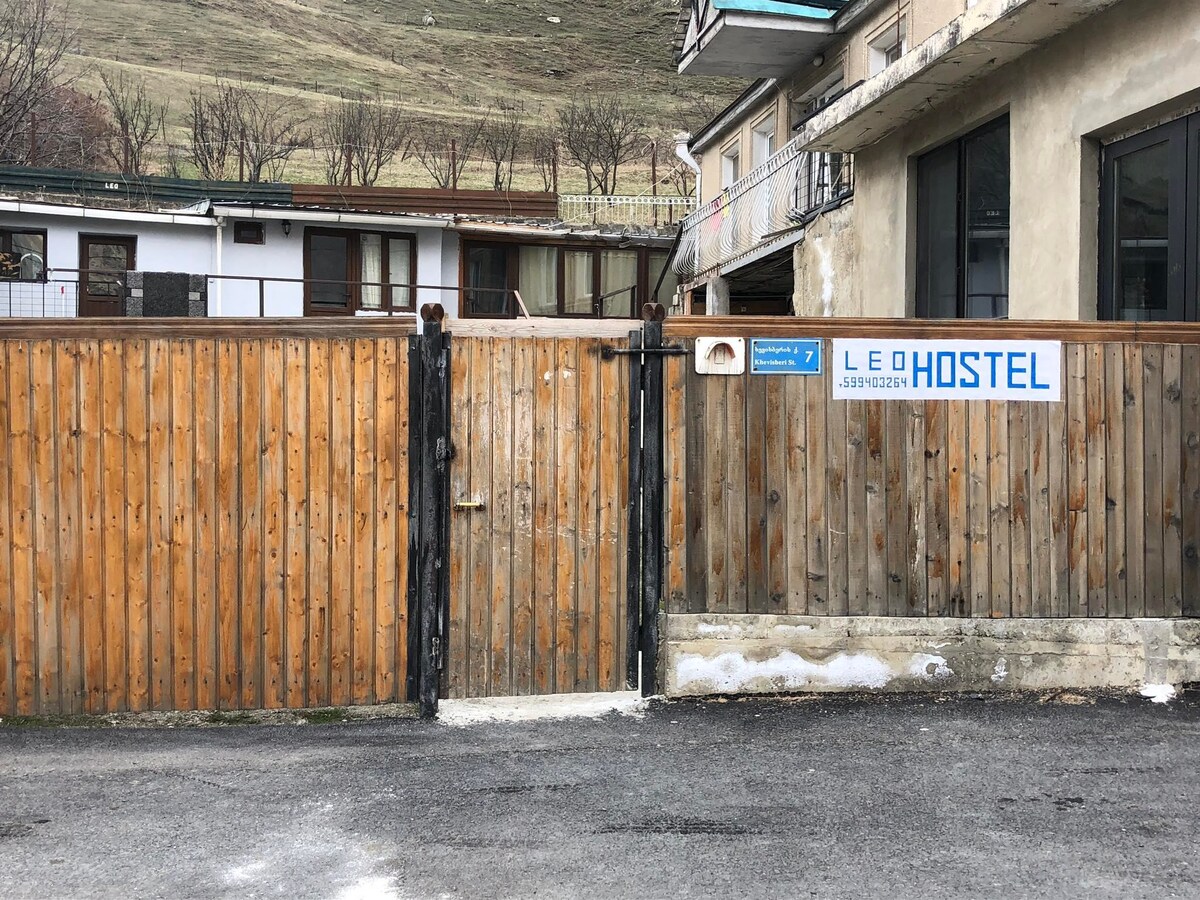 A wooden gate marks the entrance to Leo Hostel, with a sign displaying the hostel name affixed to the fence. The surroundings feature a paved area with a view of the hills in the distance, creating a serene atmosphere.
