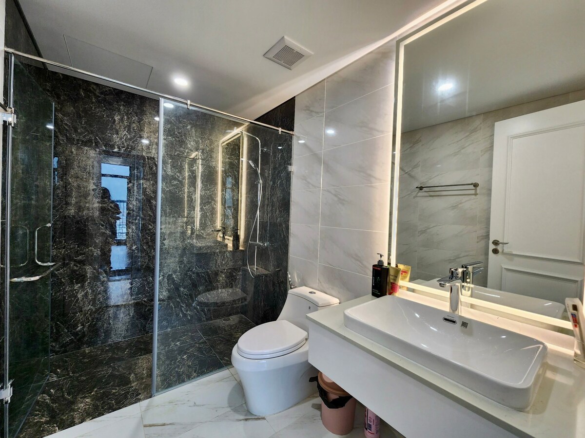 A modern bathroom showcases elegant marble finishes and a glass-enclosed shower. A sleek toilet is positioned beside a spacious vanity with a large sink and illuminated mirror, reflecting a clean and contemporary design. Neutral tones and bright lighting create a fresh ambiance.