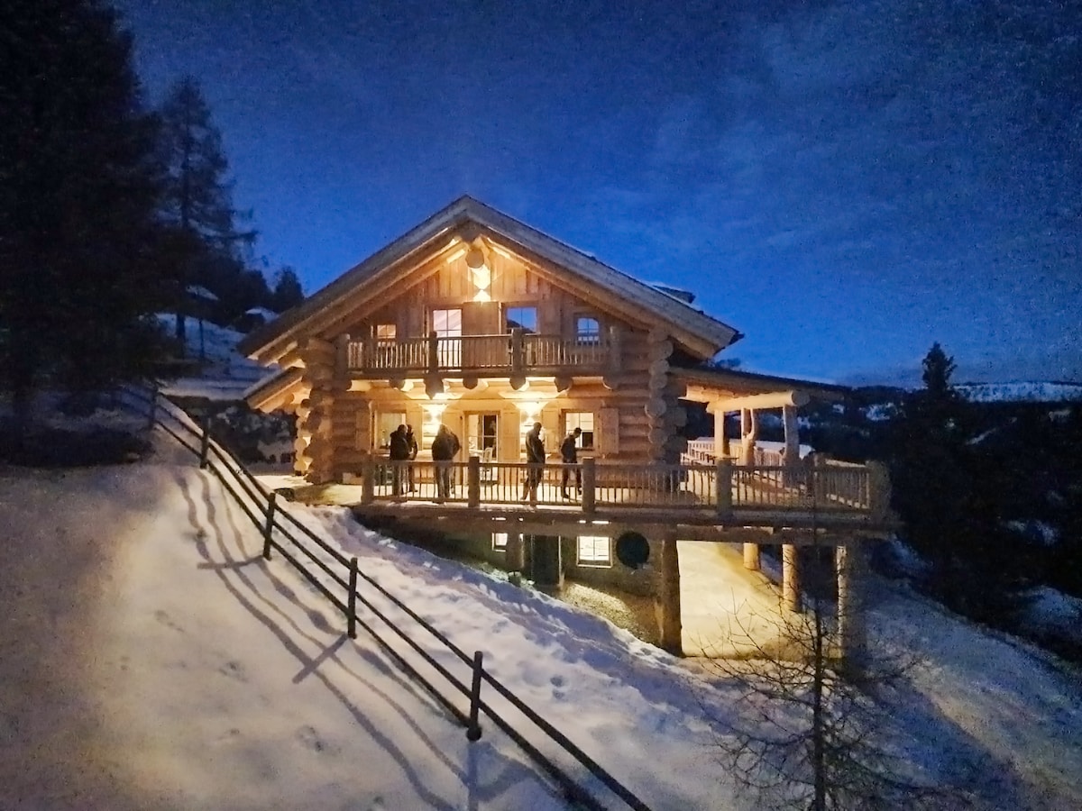 The round timber house is illuminated against a night sky, highlighting its spacious exterior deck. Snow blankets the surrounding landscape, with gentle slopes leading to the entrance. Warm lighting enhances the welcoming appearance of the structure, inviting guests to enjoy the outdoor space.