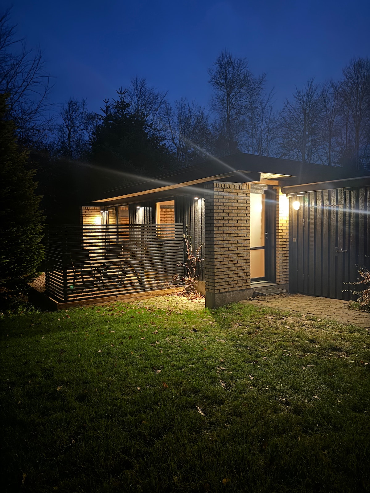 The exterior of a newly renovated annex is shown at dusk, featuring a modern brick facade and well-lit entrance. Surrounding greenery is visible, and a wooden deck with slatted design offers a welcoming space. Soft lighting creates a calm ambiance around the entrance.