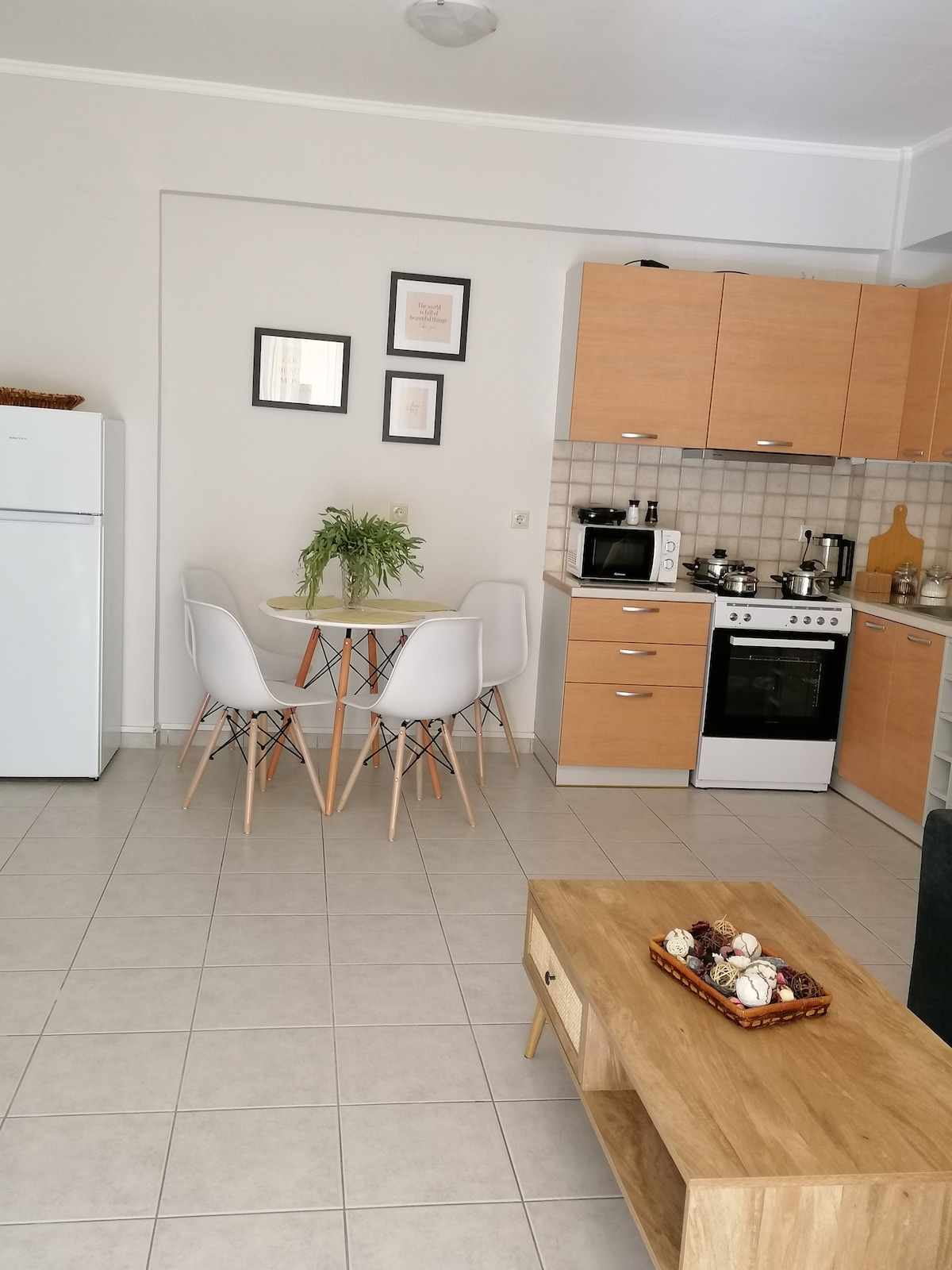 A modern kitchen is equipped with essential appliances, including a refrigerator and an oven. A round dining table with four white chairs is positioned near the walls, alongside wooden cabinetry. Decorative elements, such as a potted plant and a coffee table with an arrangement of items, enhance the space.