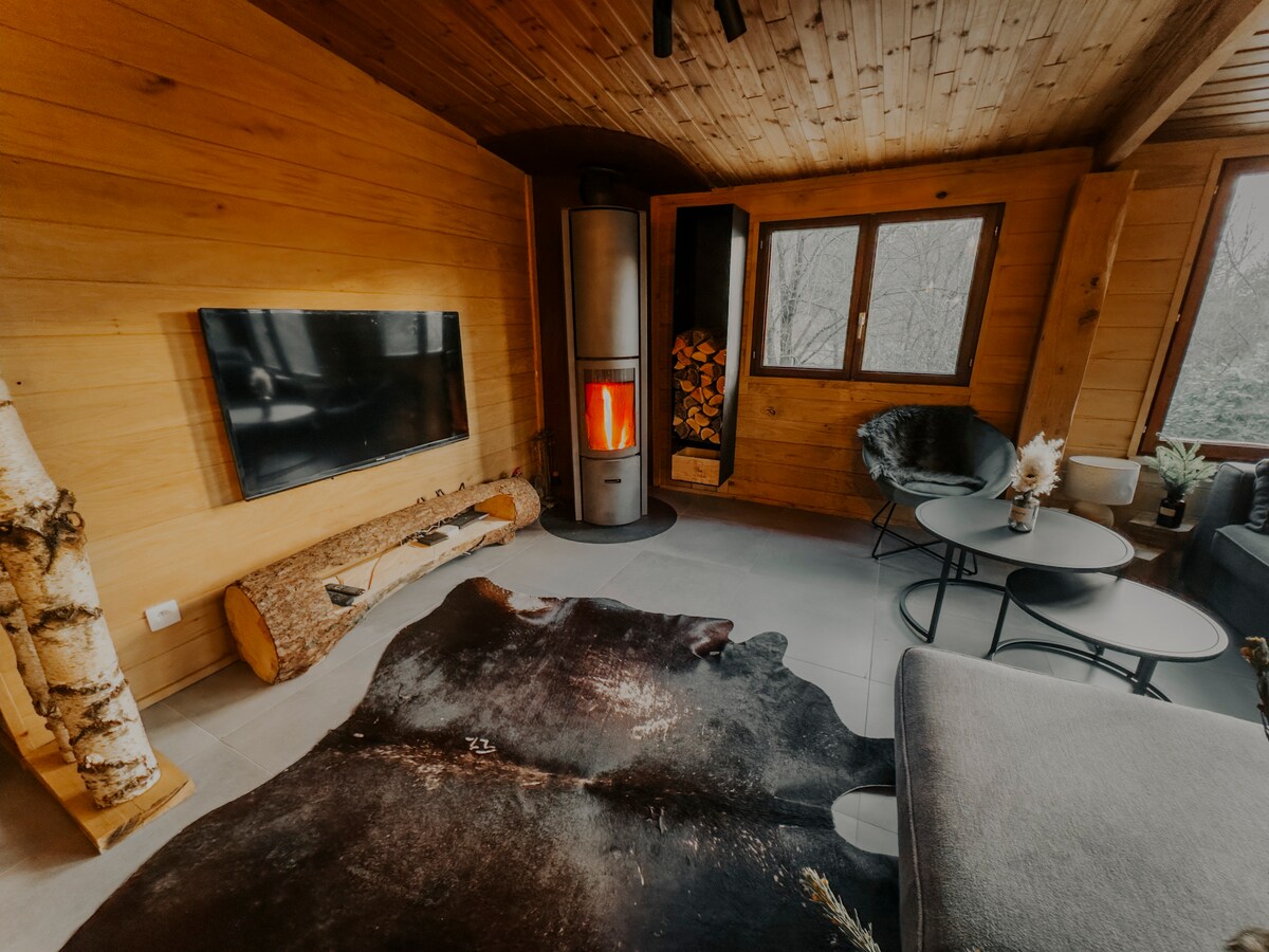 A cozy living area features wooden walls and a modern fireplace with stacked logs. A flat-screen TV is mounted on the wall, and a large cowhide rug lies on the floor. Comfortable seating includes two black chairs and two side tables, inviting relaxation.