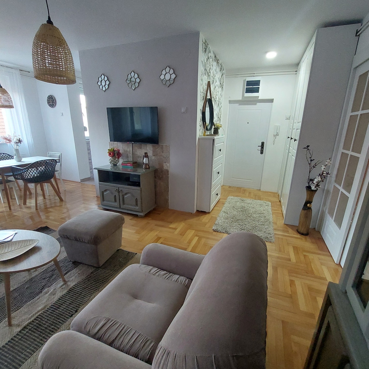 A spacious living area features comfortable seating and a decorative coffee table. A television is mounted on the wall above a stylish sideboard. Large windows allow natural light to brighten the space, enhancing the warm atmosphere created by neutral tones and textured decor.
