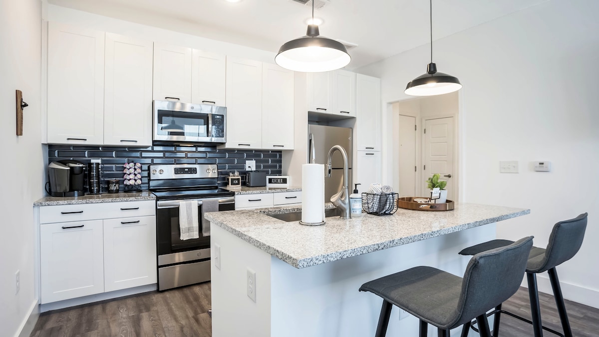 A modern kitchen features sleek white cabinetry and a dark backsplash. Stainless steel appliances, including a refrigerator and stove, are visible. A spacious island with a granite countertop is equipped with two black bar stools and is adorned with a small plant and kitchen essentials.