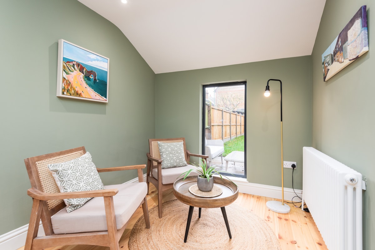 A tranquil seating area features two wooden chairs with soft cushions, positioned around a round coffee table. A small potted plant adds a touch of greenery. Natural light enters through a sliding door, leading to the private garden, while artwork decorates the walls.
