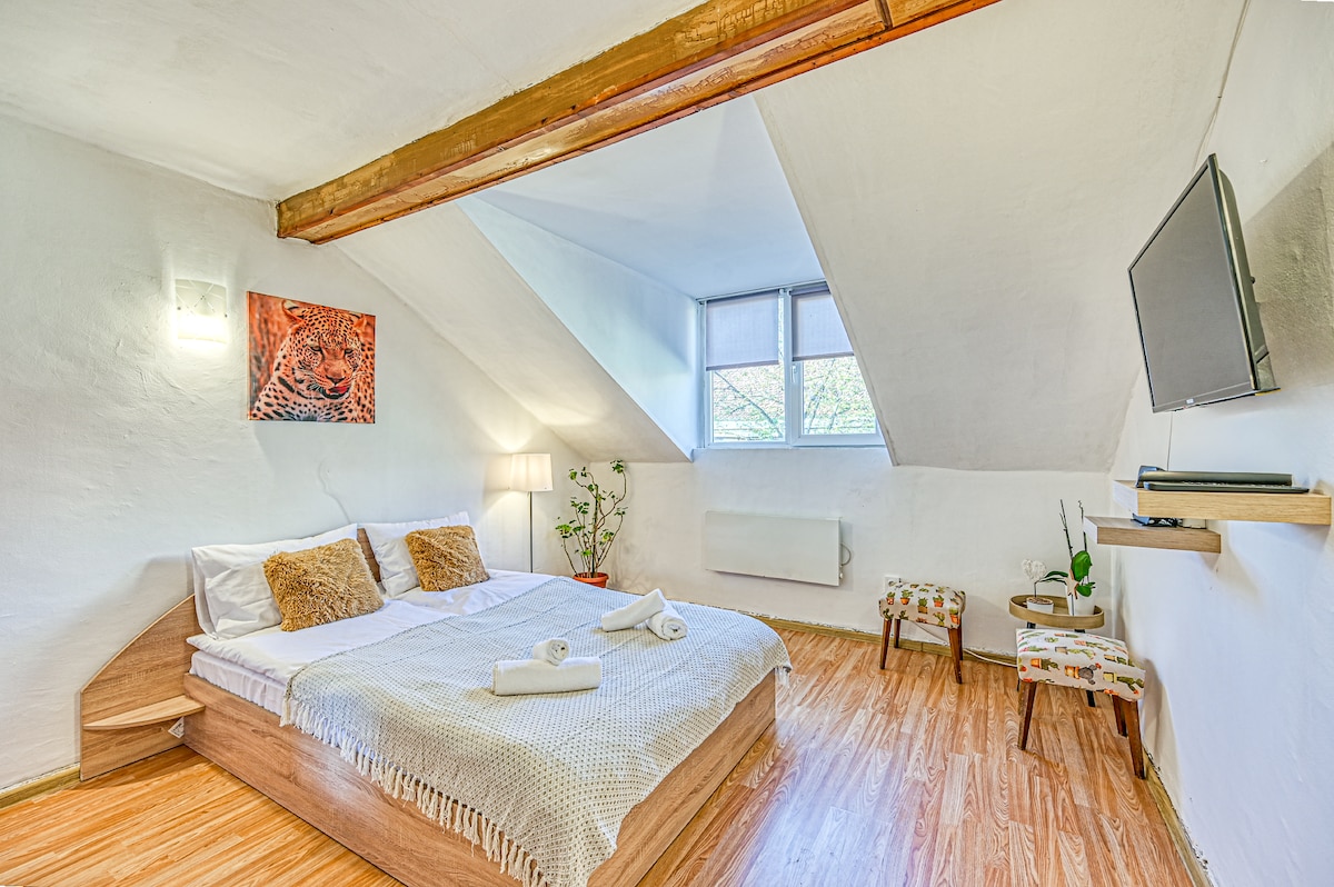 A spacious sleeping area features a comfortable bed with a light-colored knitted throw and neatly arranged towels. Natural light flows through two windows, highlighting the warm wooden flooring. A cozy lamp and a decorative artwork add subtle charm, while a small TV is mounted on the wall.