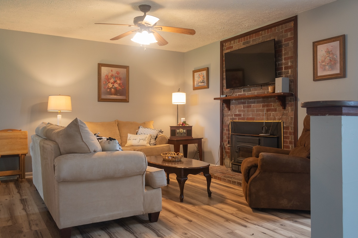 A comfortable living room features a light-colored sectional sofa and a recliner, both arranged around a wooden coffee table. A brick fireplace is positioned beneath a wall-mounted television. Soft lighting from a lamp and overhead fan creates a warm atmosphere.