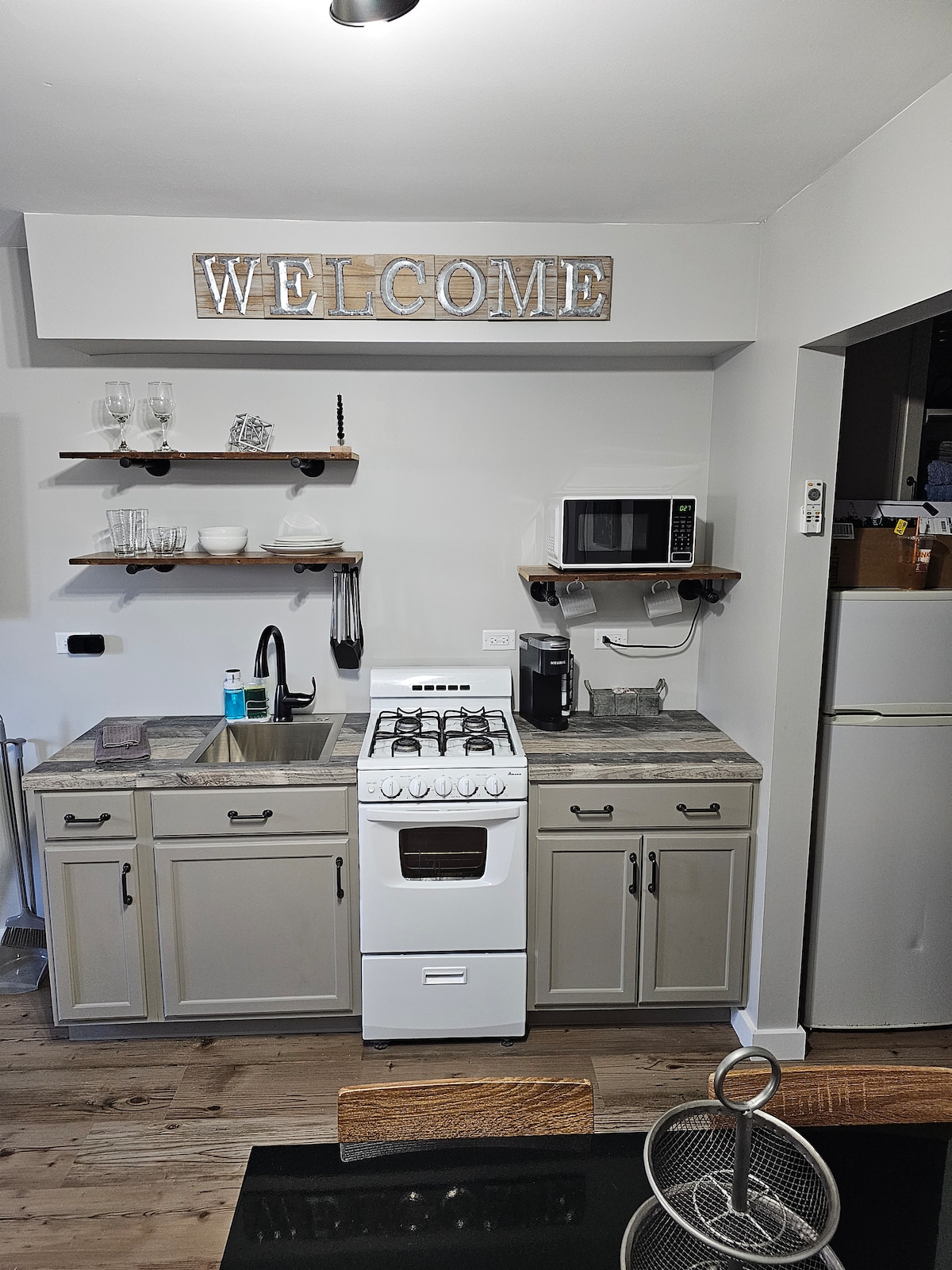 A compact kitchenette features a gas stove, microwave, and refrigerator, along with ample countertop space. Open shelving displays dishware and glassware, while a welcome sign adds a friendly touch to the space. The muted color palette contributes to a cozy atmosphere.