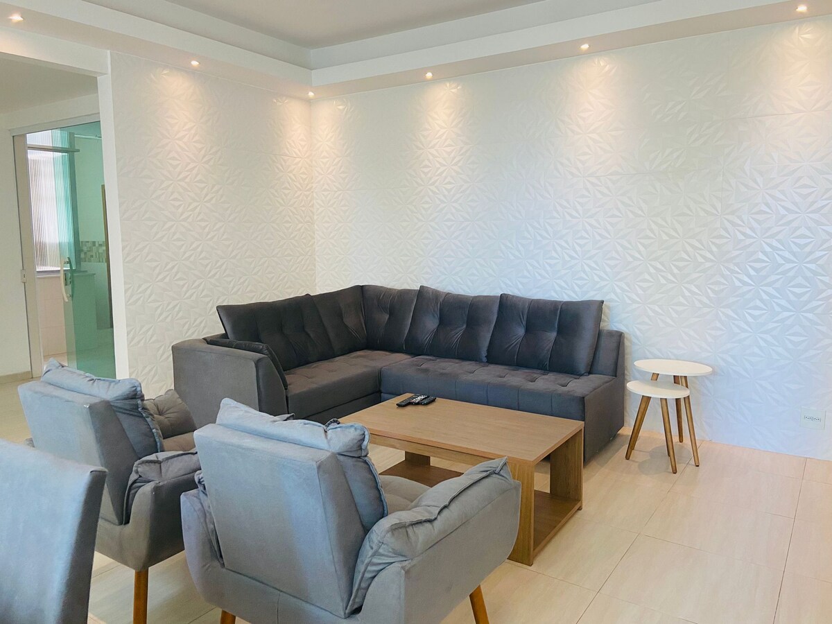 A spacious living area is furnished with a grey sectional sofa and two armchairs, arranged around a light wood coffee table. Soft lighting highlights the textured wall design, enhancing the modern and clean atmosphere.