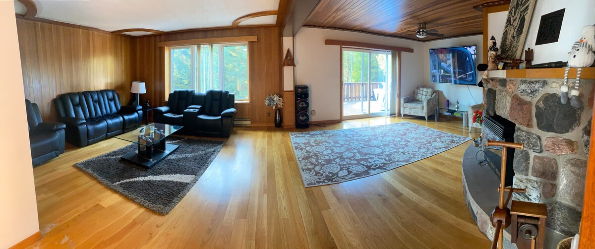 A spacious living area features large windows allowing natural light to illuminate the hardwood floors. Black leather sofas are arranged around a glass-topped coffee table, with a large area rug adding texture. A stone fireplace and a sliding door leading to a deck complement the inviting space.