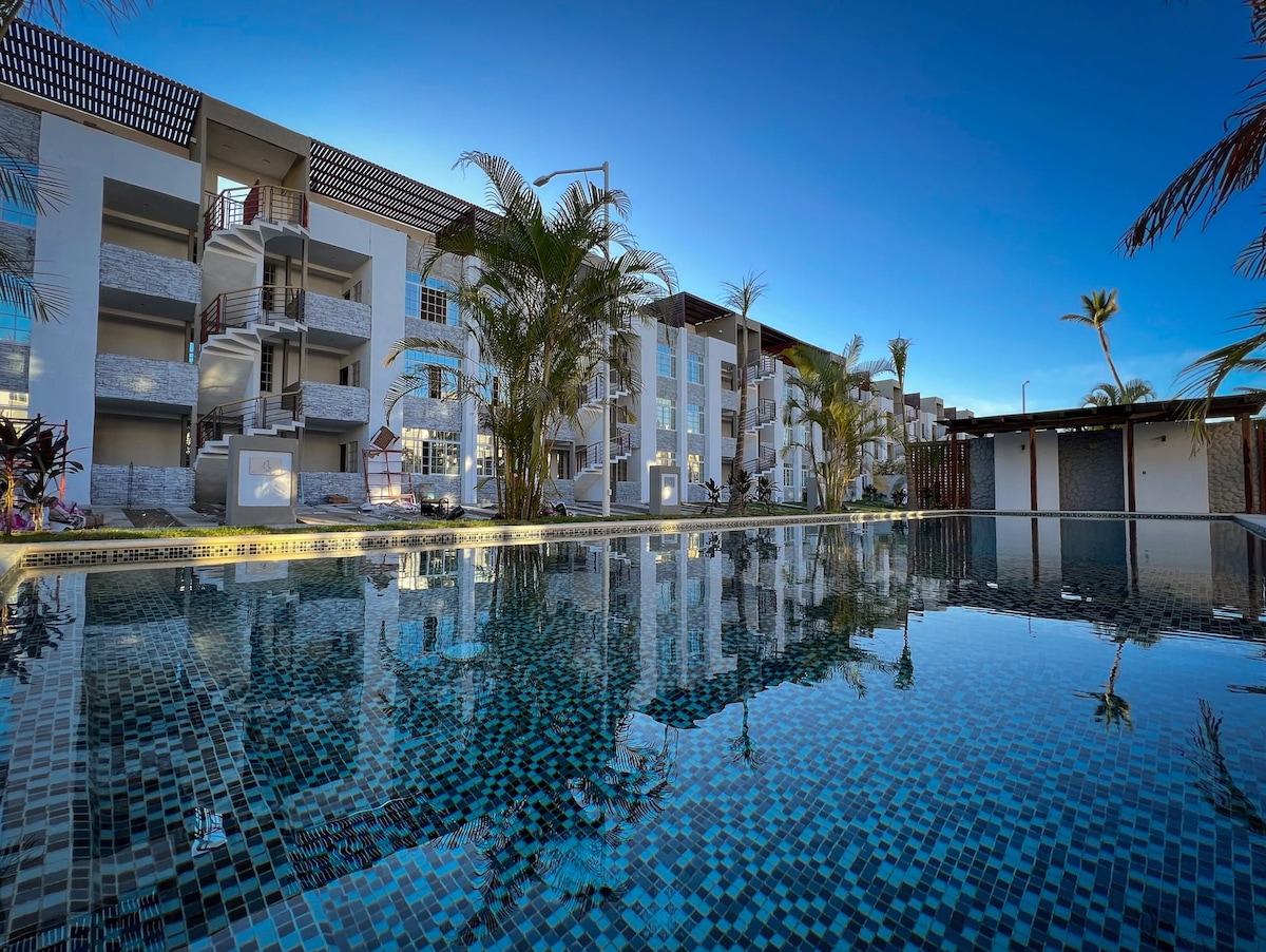 The image displays a modern apartment complex beside a large swimming pool. Palm trees are positioned around the pool area, and the evening sky adds a serene ambiance. The building features multiple stories with glass accents reflecting in the water.