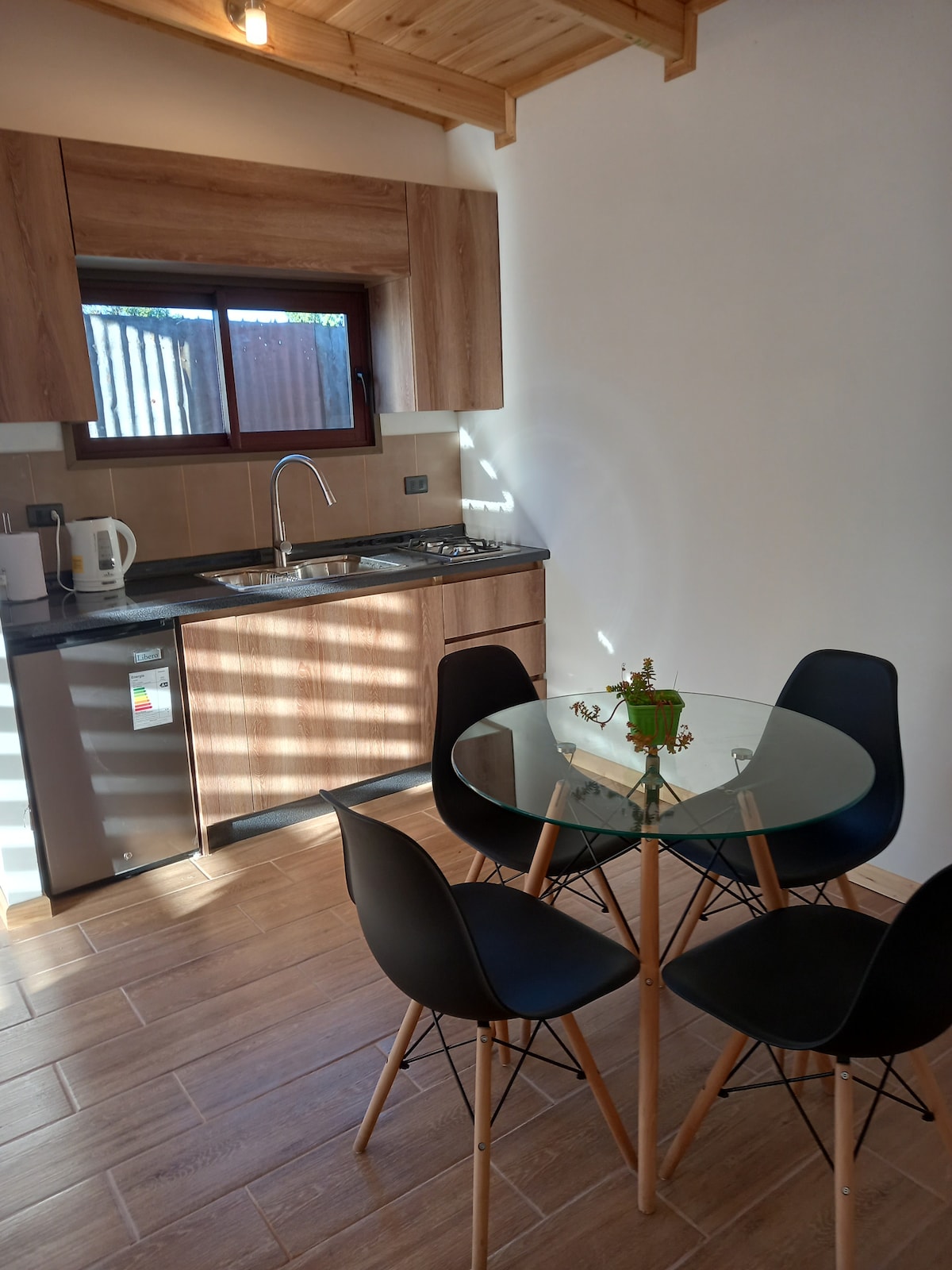 A modern kitchen area is shown with polished wood accents and a large window allowing natural light. A round glass table with four black chairs is positioned at the center. Kitchen fixtures and appliances are organized along the counter, providing a functional space.