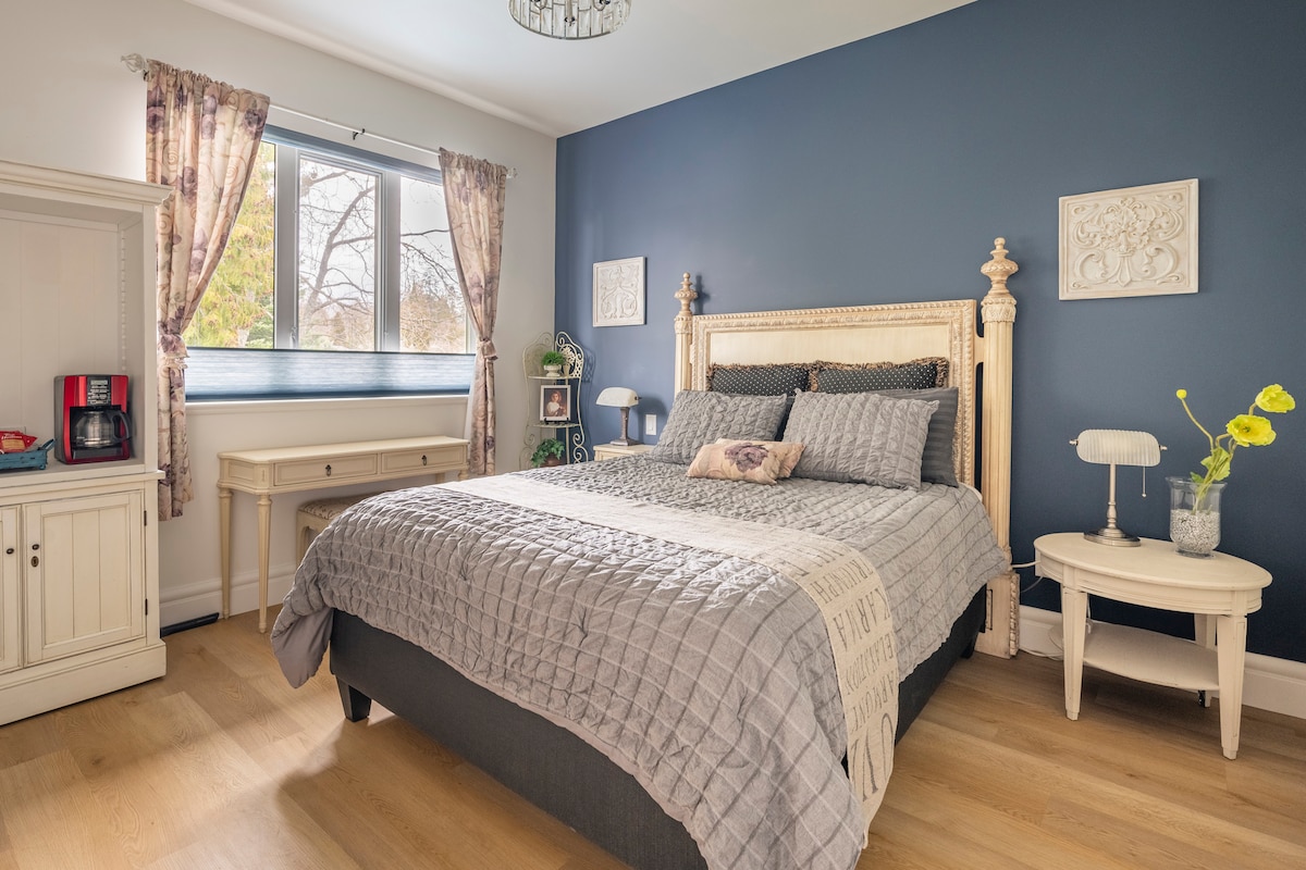 A serene bedroom is enhanced by a deep blue accent wall and large window that invites natural light. A queen size bed is adorned with a textured blanket, flanked by two nightstands. A small table with flowers adds a touch of elegance to the space.