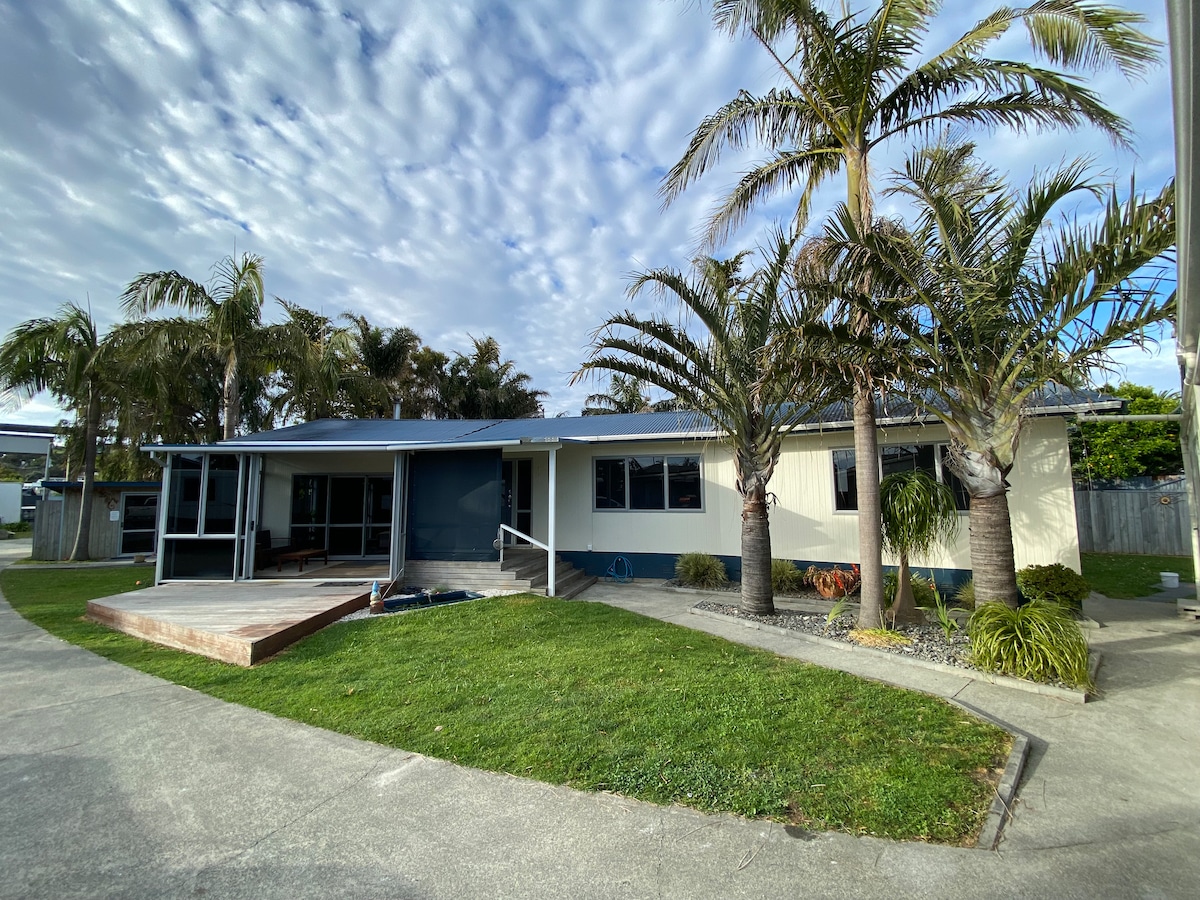 A single-story building is framed by several palm trees, with a grass lawn in the foreground. A wooden deck leads to the entrance, which features a stairway that provides access to the wide porch. The sky above is partly cloudy.