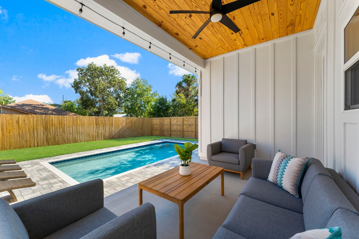 A comfortable outdoor seating area features modern gray sofas arranged around a wooden coffee table. In the background, a private heated pool is visible, surrounded by a neat lawn and enclosed by wooden fencing, under a clear blue sky.