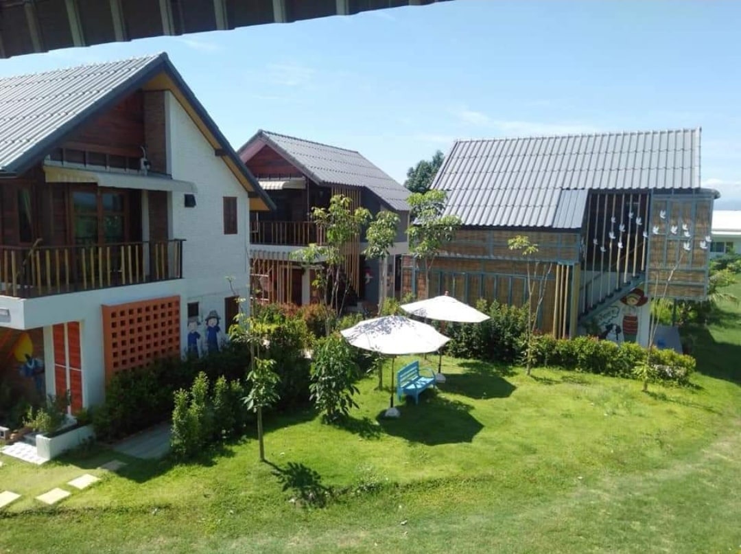 Two charming guesthouses are visible, set amid lush greenery. Each building features distinct architectural styles, with wooden accents and large windows. A shaded seating area and a park bench are situated on the manicured lawn, providing inviting spots to relax outdoors.
