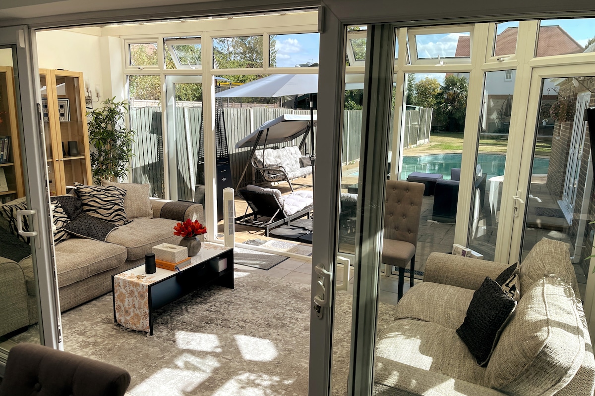 The conservatory is bathed in natural light, featuring spacious seating arrangements and modern furnishings. Large glass doors provide a view of the heated pool and landscaped garden, while a shaded outdoor seating area adds relaxation options. The tranquil setting exudes openness and comfort.
