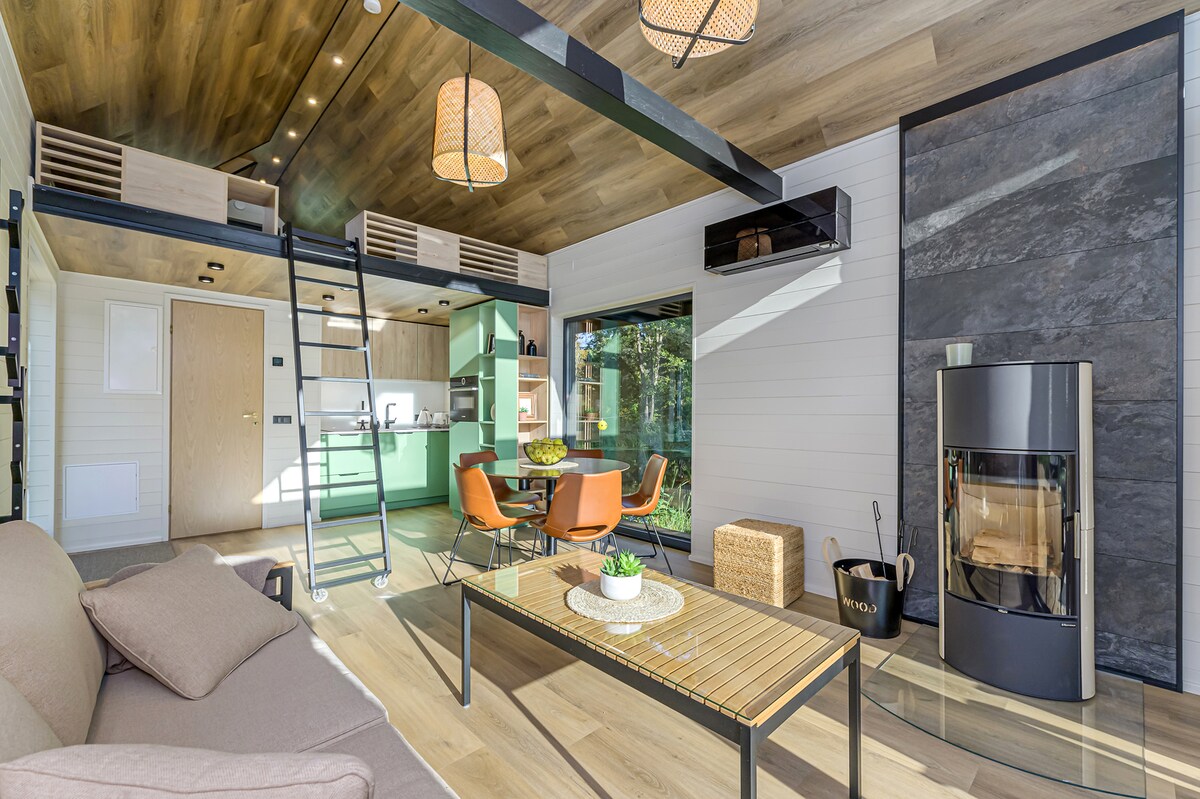 An open living space features a cozy sofa and a wooden coffee table, complemented by an elegant fireplace. A modern kitchen area is visible, with green cabinetry and a dining setup, while large windows provide views of the surrounding nature.