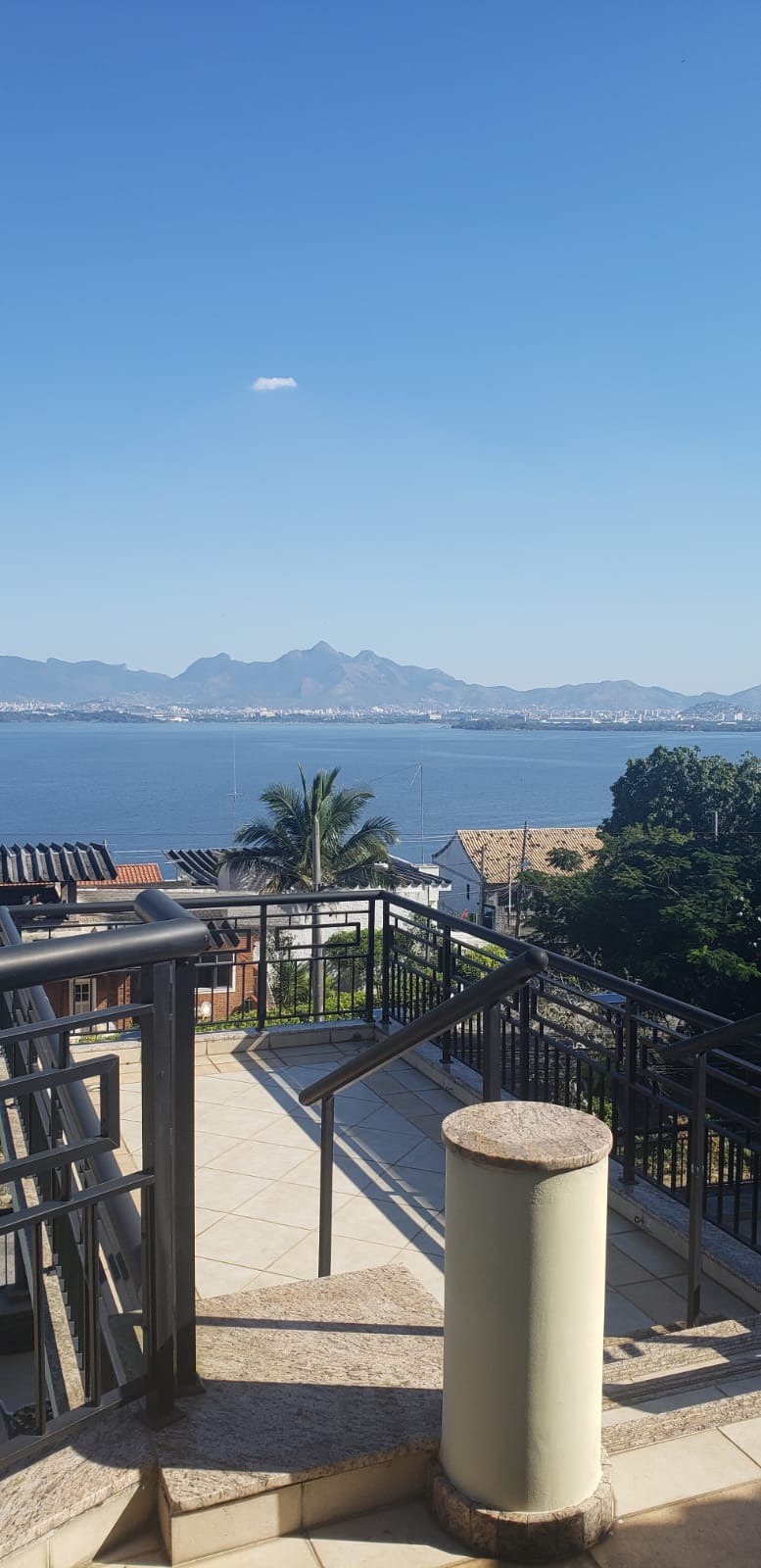 A scenic view of water and distant mountains is captured from a balcony. The outdoor terrace features textured stone flooring and a metal railing, complemented by lush greenery visible from below. The clear blue sky enhances the tranquil atmosphere of the space.
