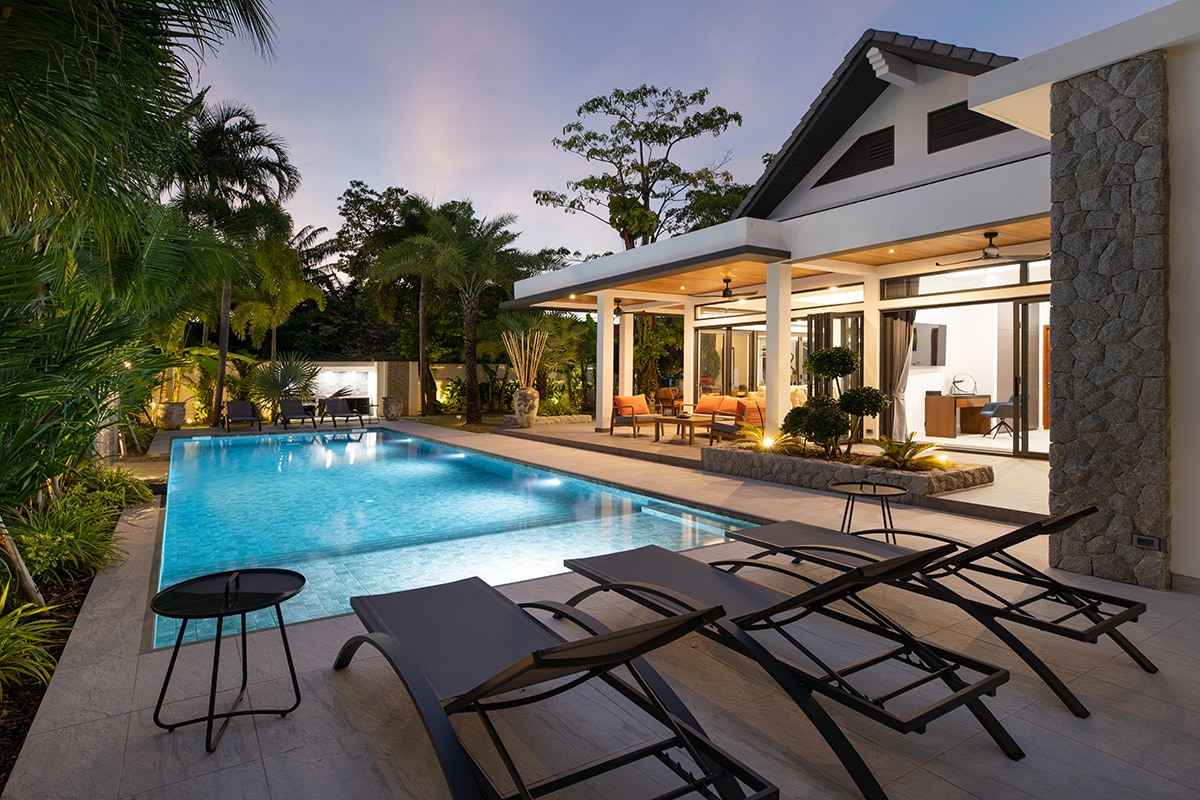 The image captures a serene swimming pool surrounded by elegant lounge chairs, with tropical greenery framing the space. A covered patio is visible in the background, featuring comfortable seating, all illuminated by soft evening light under the clear sky.