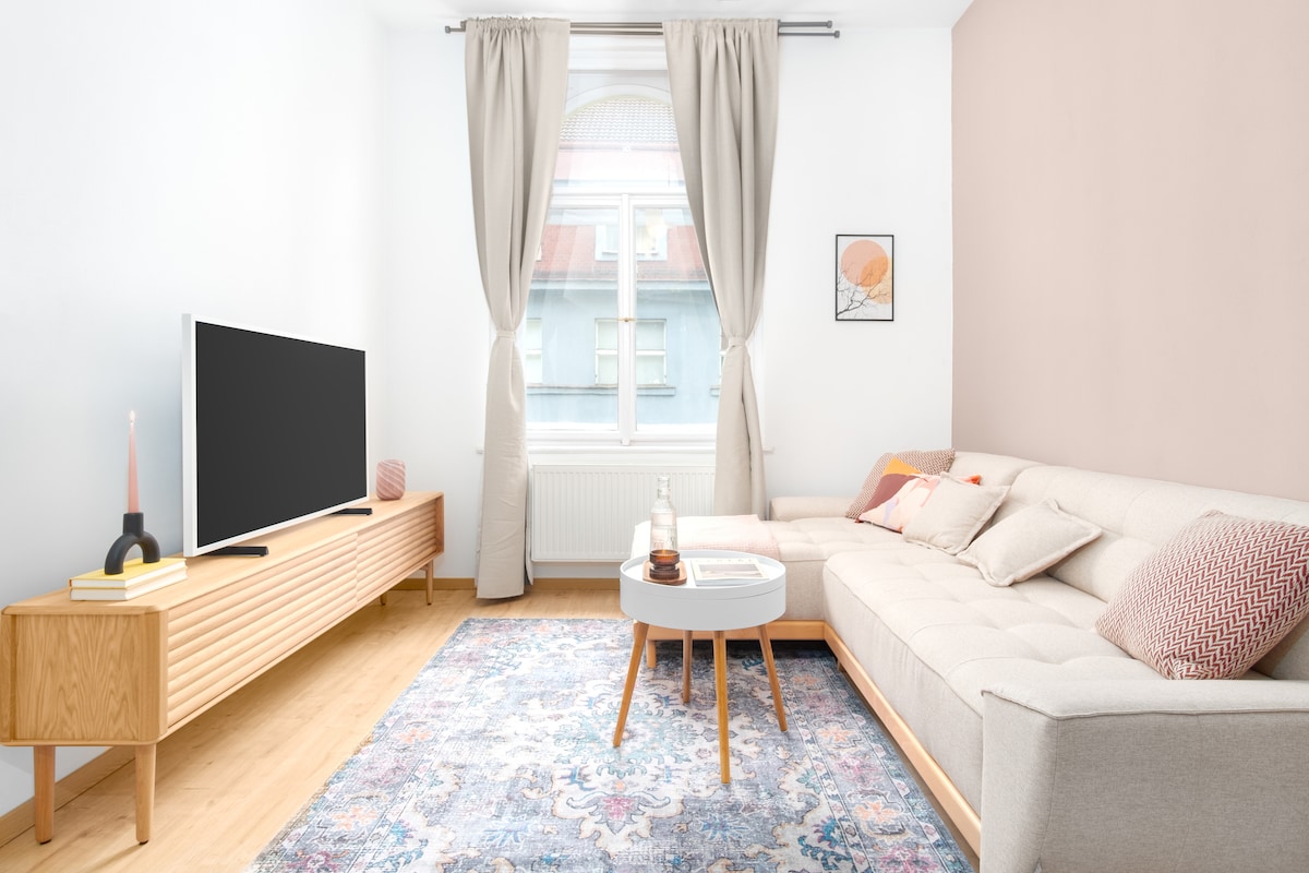 A light-filled living room features a spacious gray couch adorned with colorful cushions. A stylish coffee table sits on a patterned rug, while a wooden media console supports a large flat-screen TV. Flowing curtains frame the large window overlooking the calm street.