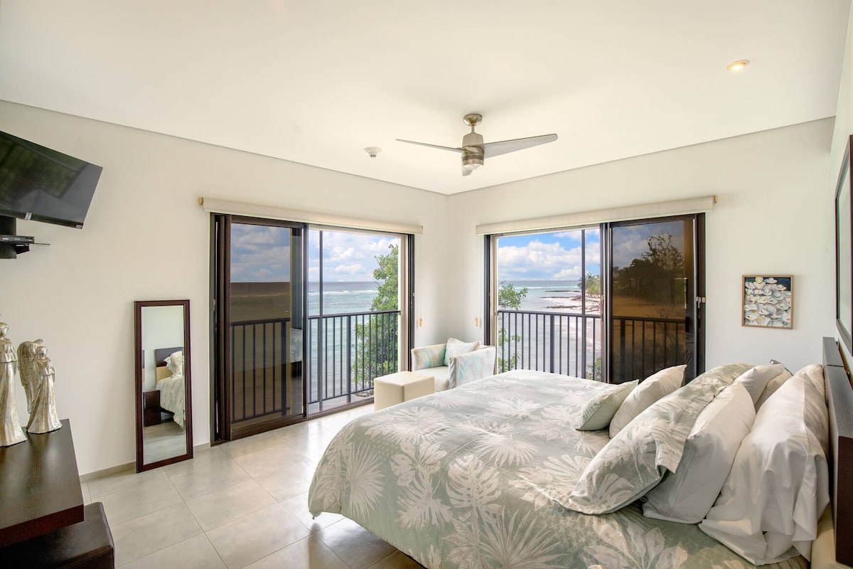 A spacious bedroom features a large bed adorned with soft linens, complemented by elegant palm-print decor. Two sliding glass doors offer stunning ocean views, while a ceiling fan provides comfort. A dresser and mirror enhance the room's functionality and style.