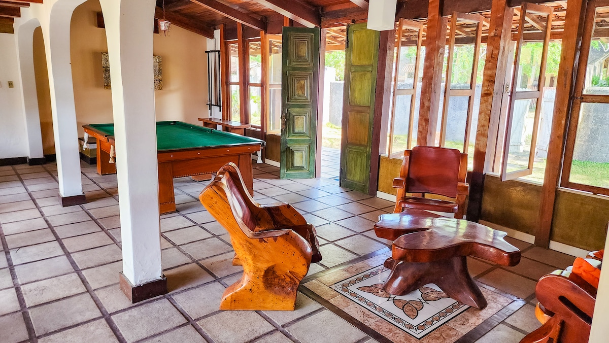 An airy living space features a wooden pool table, complemented by rustic furniture made of natural wood. Sunlight filters through large windows, illuminating the tiled floor and enhancing the cozy atmosphere. The room is designed for relaxation and socializing.