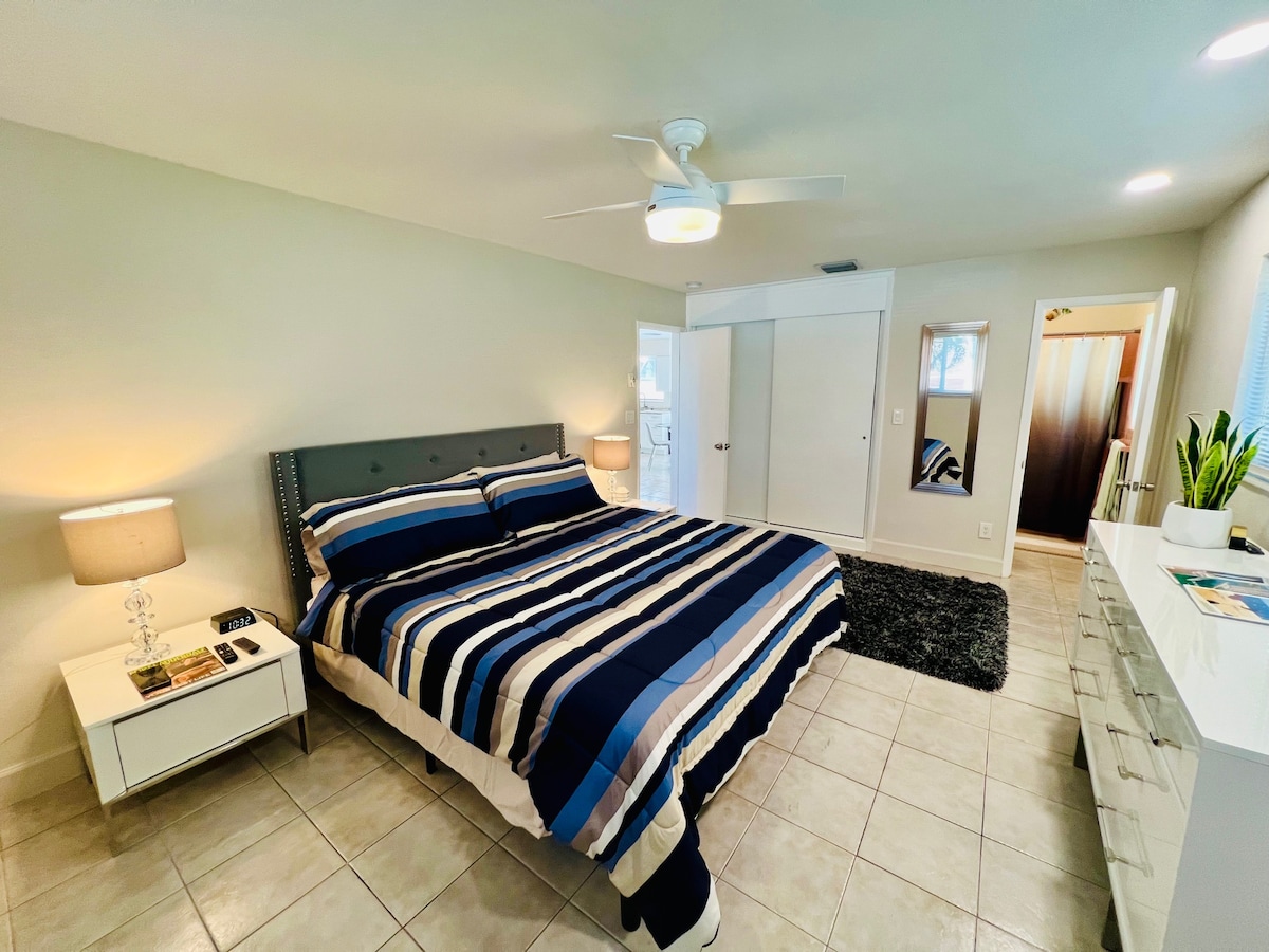 A spacious bedroom features a large bed with a striped comforter and side tables with lamps. A ceiling fan is visible, and a dresser is set against a wall. Light floods in from the adjacent doorway, while tile flooring adds a clean touch.