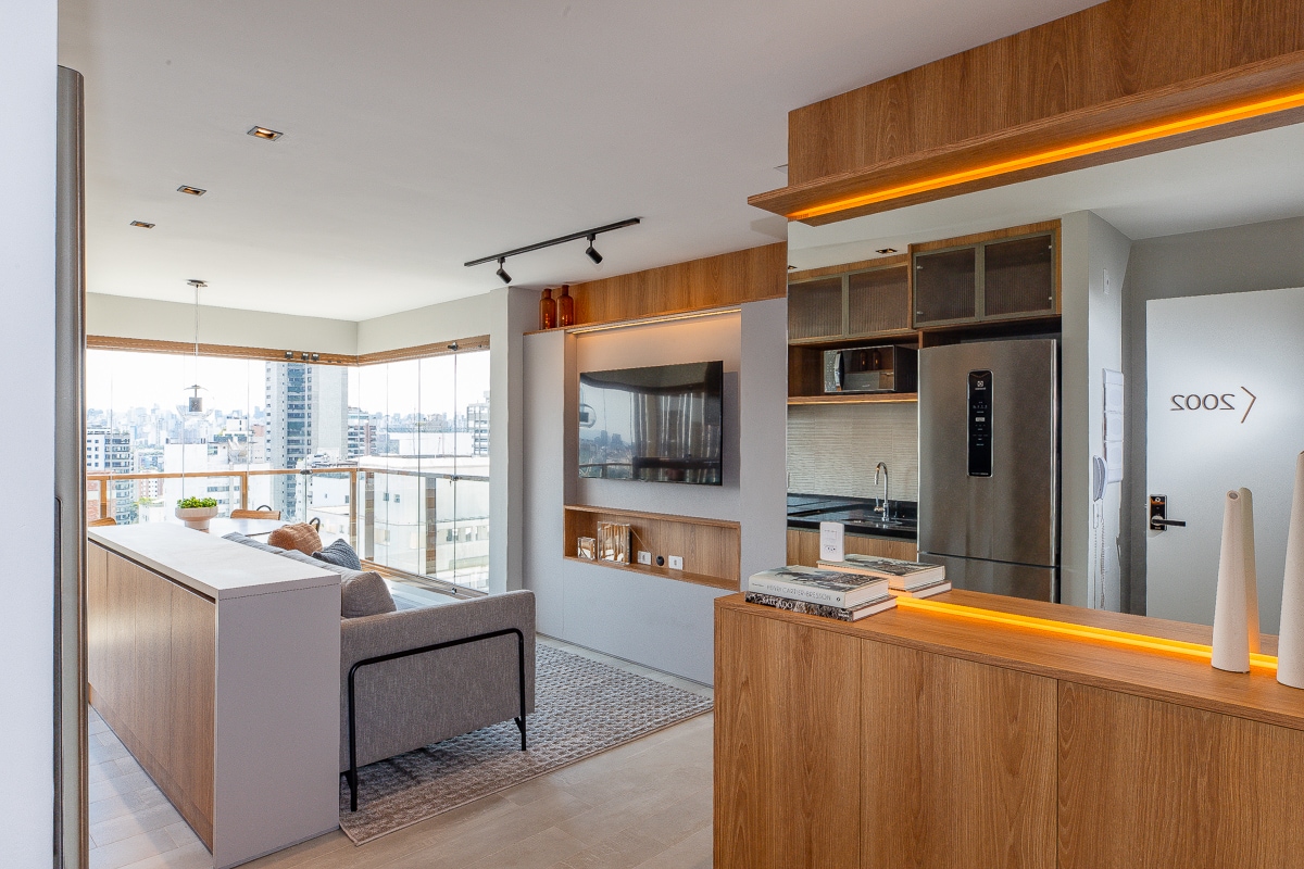 An open living space is presented, featuring a cozy sofa and a sleek media unit against a wooden accent wall. A modern kitchen with stainless steel appliances is visible, complemented by a dining area. Natural light flows through large windows, enhancing the airy atmosphere.