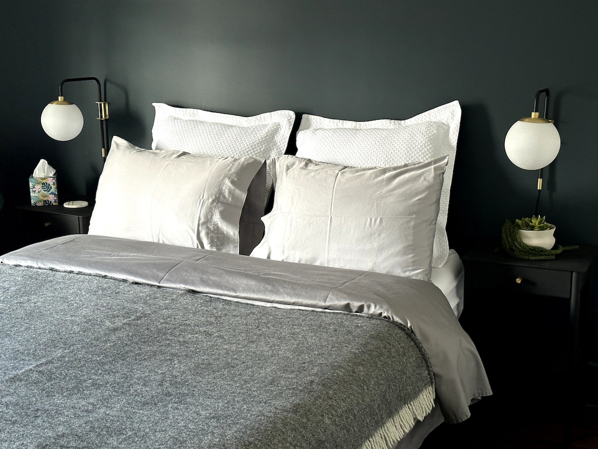 A neatly arranged sleeping area features a spacious bed with soft, muted bedding against a dark wall. Two bedside lamps provide gentle illumination, and a small plant adds a touch of greenery on a nearby nightstand.