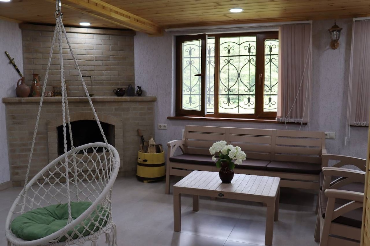 A welcoming living area features a hanging swing chair and a wooden coffee table. Natural light enters through a large window adorned with decorative ironwork. A stone fireplace adds a cozy touch, while potted plants enhance the relaxing atmosphere.
