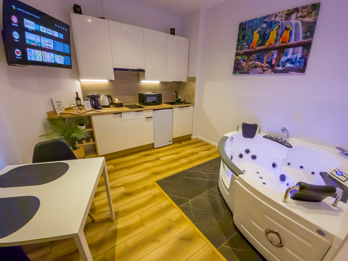An open-plan space features a modern kitchen with white cabinetry, a dining table for two, and a wall-mounted television. A hydro massage bathtub is positioned in the corner, surrounded by dark flooring and warm wood accents, providing a relaxing ambiance.
