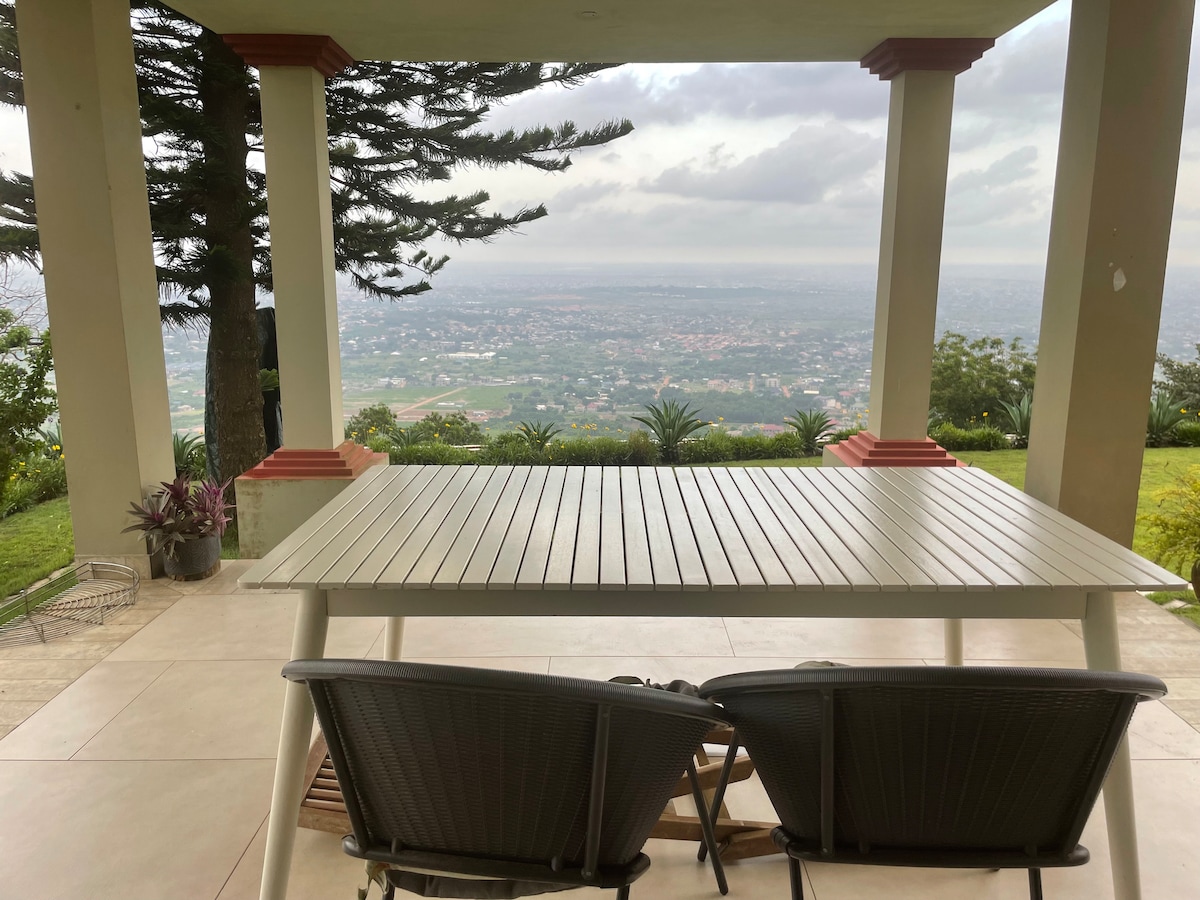 A spacious outdoor terrace is shown, featuring a large table with two comfortable chairs overlooking a sprawling view of the landscape below. Trees and plants are positioned nearby, enhancing the connection to nature.