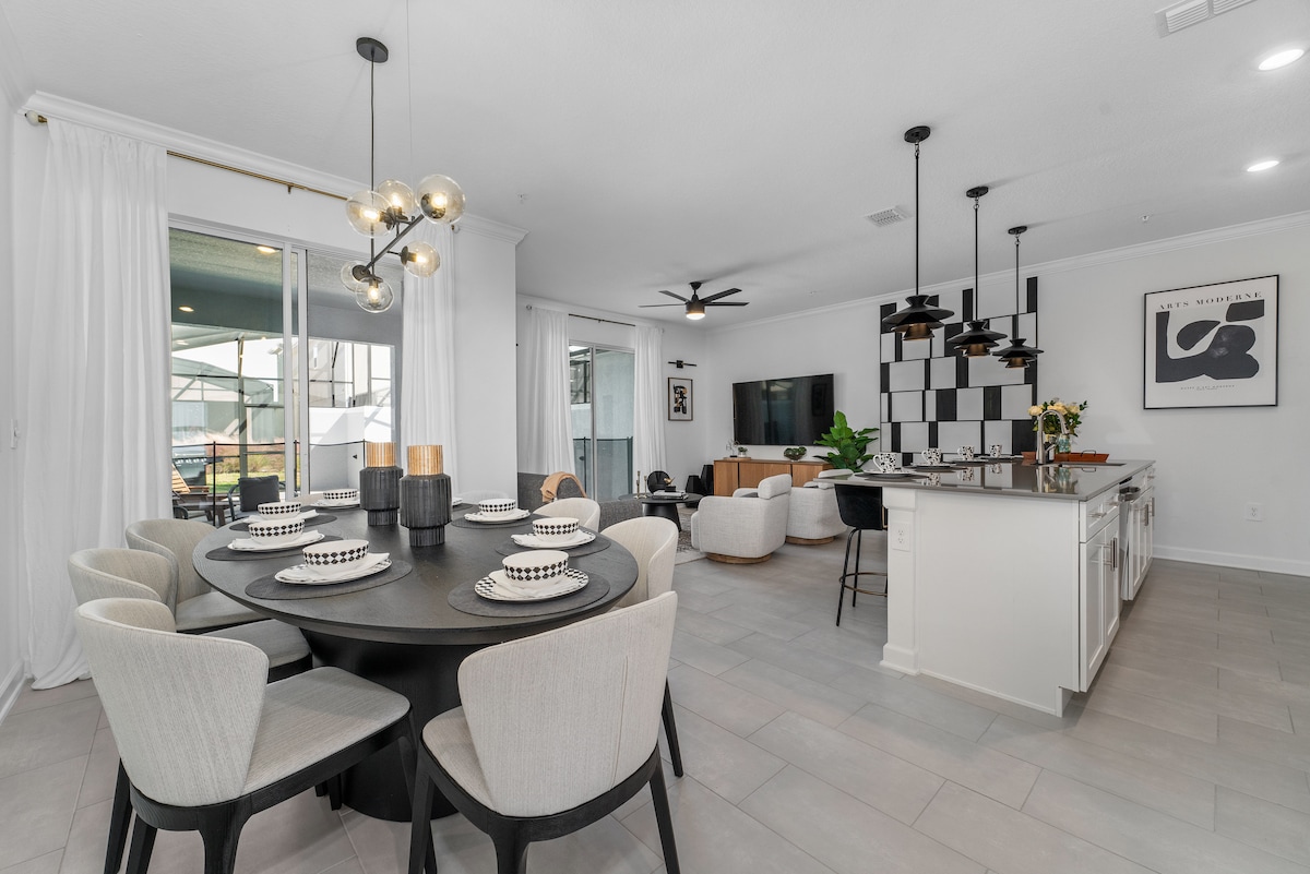 An open concept living space features a dining area with a round table set for six, surrounded by soft chairs. A modern kitchen with a large island is visible, alongside a comfortable seating area equipped with a sleek television and large windows that bring in natural light.